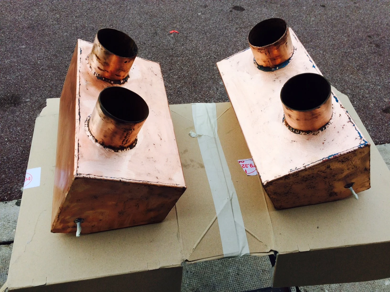 Two copper-colored metal electric heaters with black openings, placed on cardboard boxes, outdoors on asphalt surface.