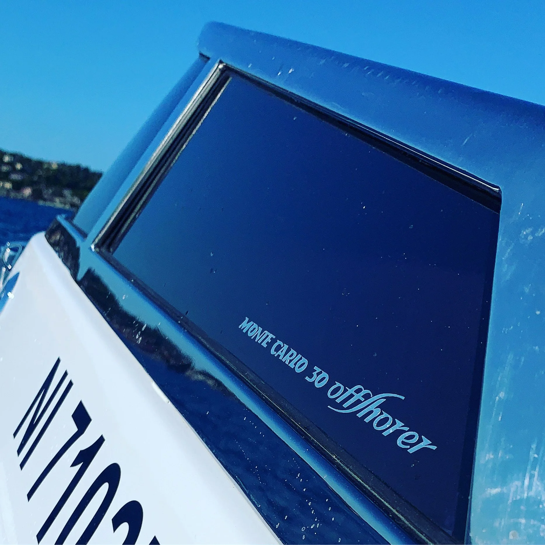 Close-up of a sign with the text 'Monte Carlo 30 Offshore' on a blue background, with a body of water and a distant shoreline visible in the background.