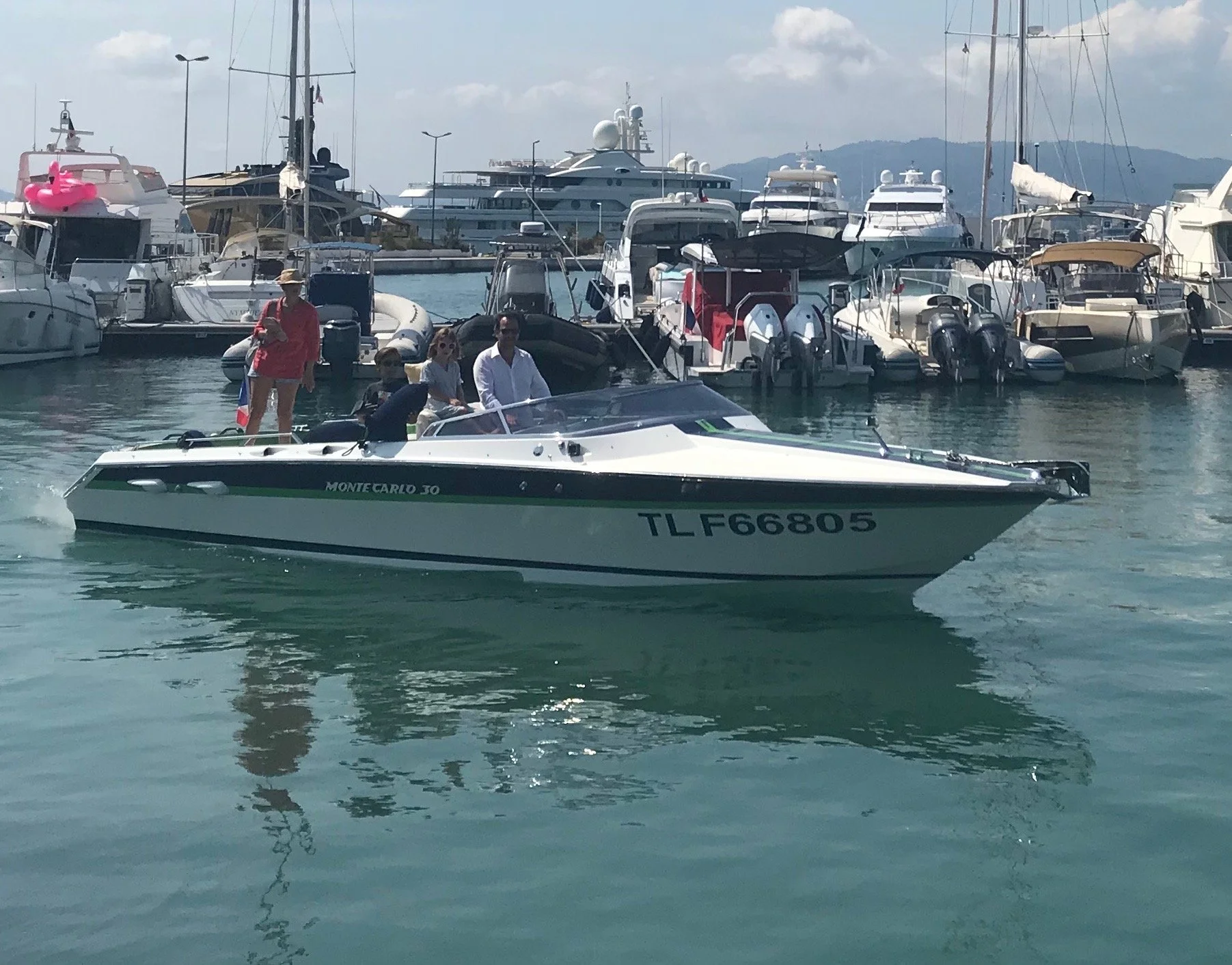 People riding a white speedboat named Monte Carlo 30 with the registration TLF66805 on a marina with multiple yachts and boats in the background.