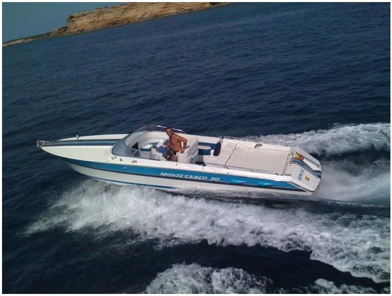 A man on a white and blue Monte Carlo JO speedboat cruising on the water near a coastline with cliffs.