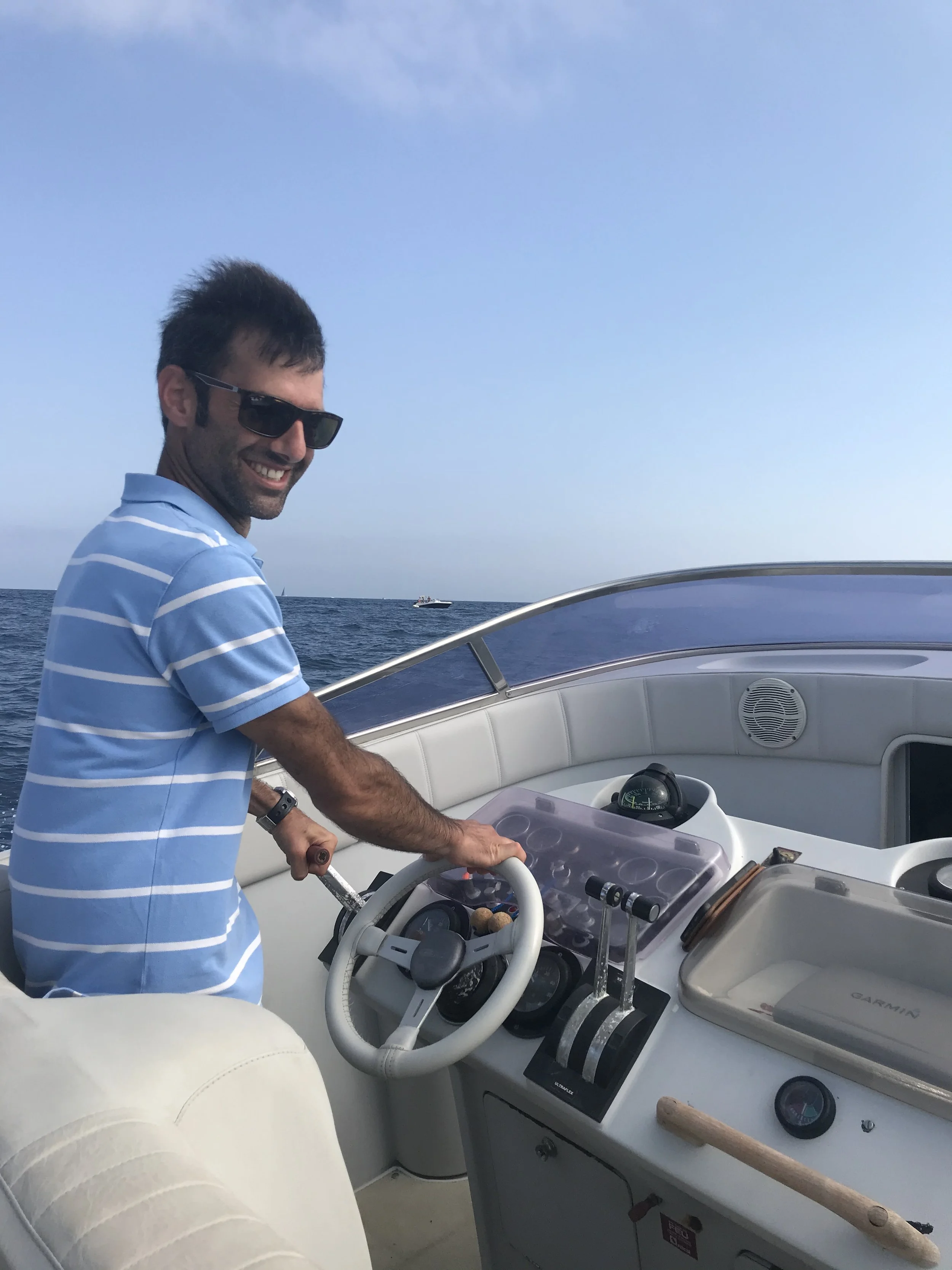 Man with sunglasses smiling while steering a boat on the ocean, wearing a light blue and white striped shirt, with a clear blue sky overhead.