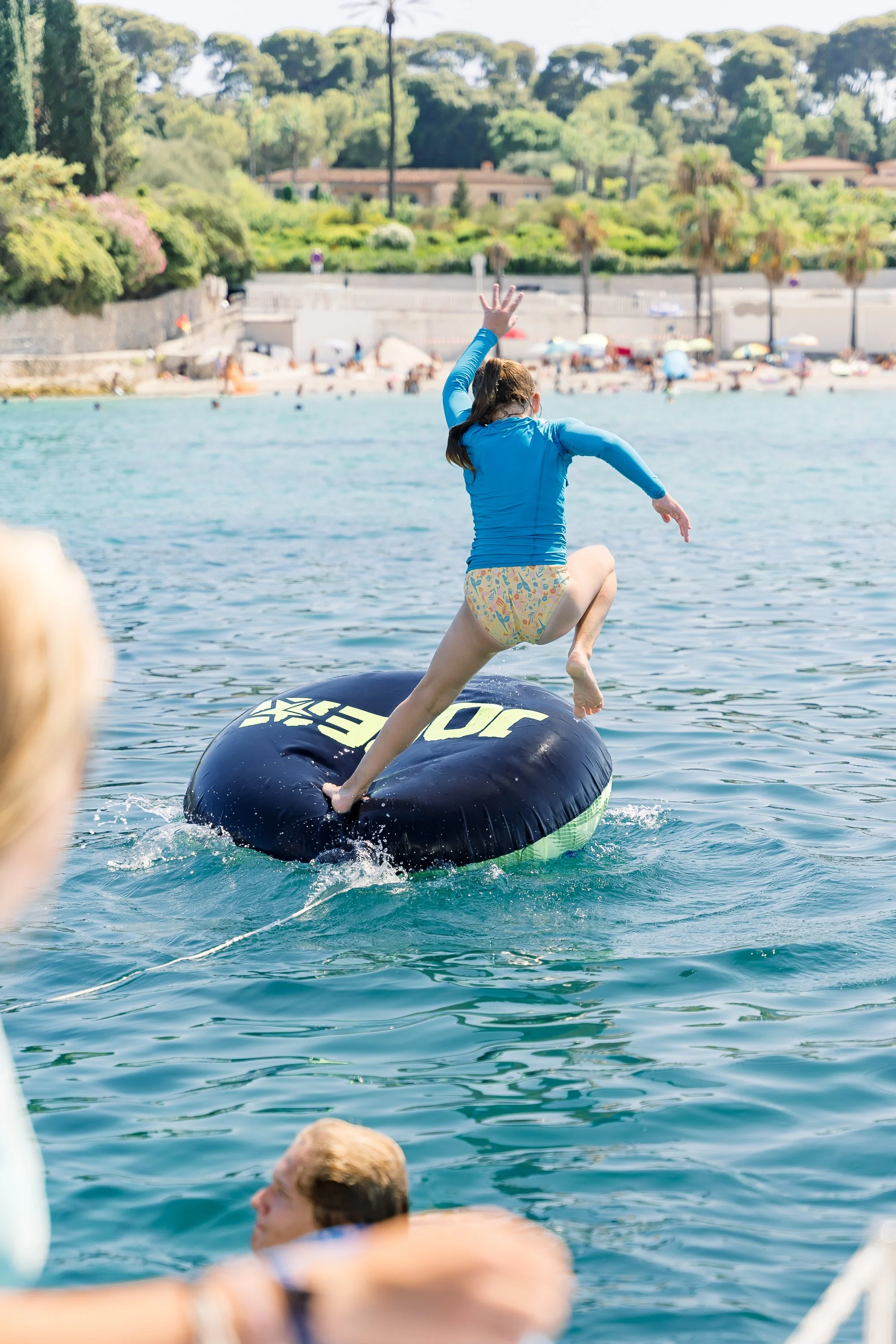 A girl in a blue rash guard and colorful swim bottoms jumps off a black inflatable water float in a body of water, with a beach and green trees in the background.