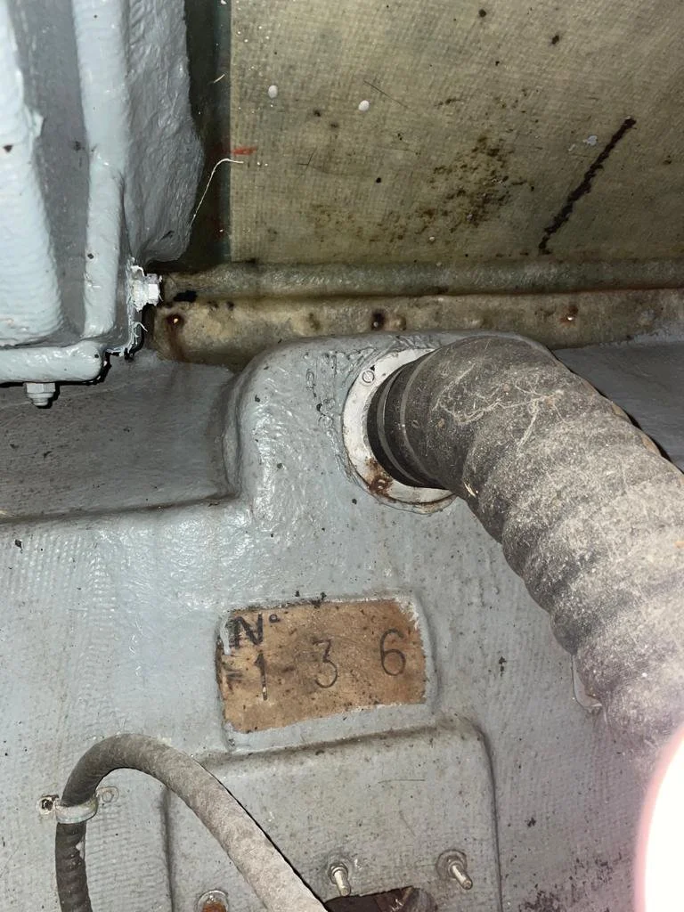 Close-up of a machine or engine with a pipe, a metal plate with handwritten numbers, and some wires and bolts, showing signs of rust and dirt.