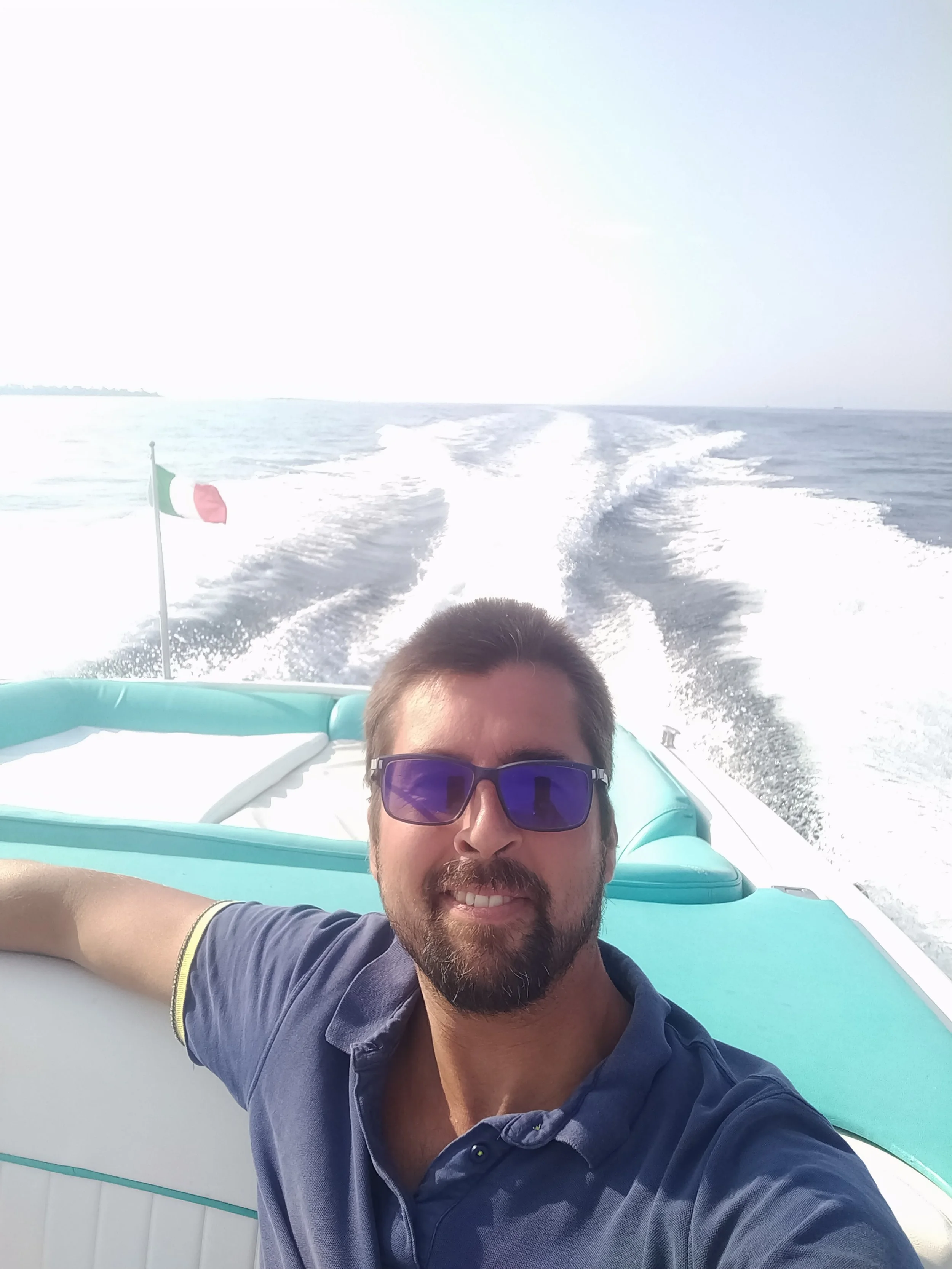 A man with sunglasses taking a selfie on a boat in the ocean, with a trail of water behind the boat and the Italian flag visible on the boat.