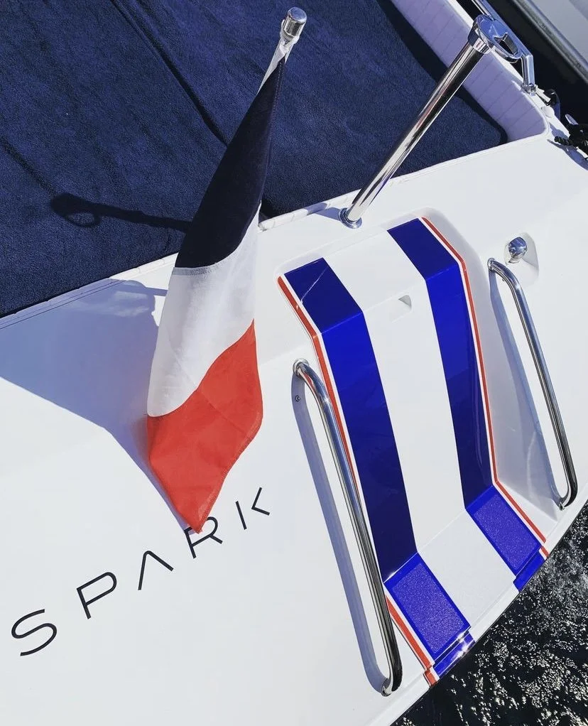 A close-up photo of a boat with a French flag and racing stripes in blue, white, and red. The word 'SPARK' is partially visible on the boat's surface.