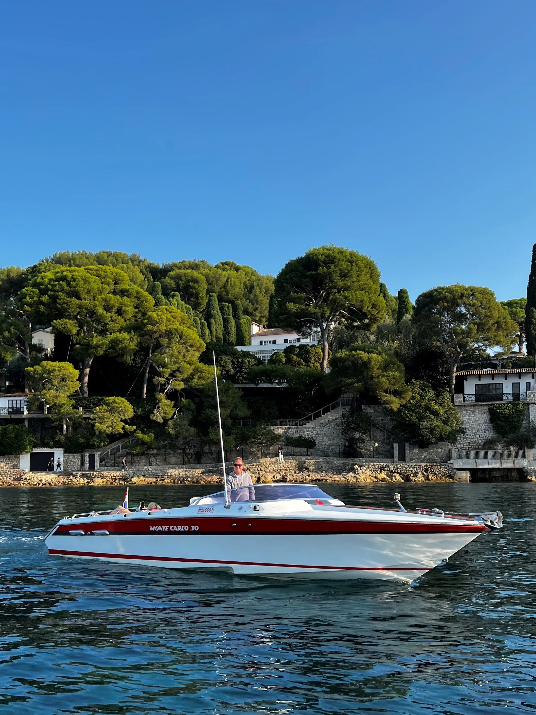 A man standing on a white and red yacht named Monte Carlo 30 on calm water with a green hillside and houses in the background under a clear blue sky.
