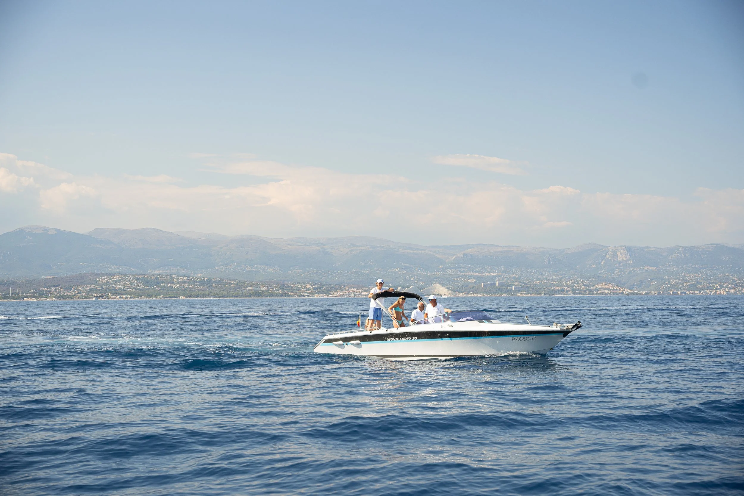 A speedboat with four people on board moving across a body of water on a sunny day, with mountains and a distant shoreline in the background.