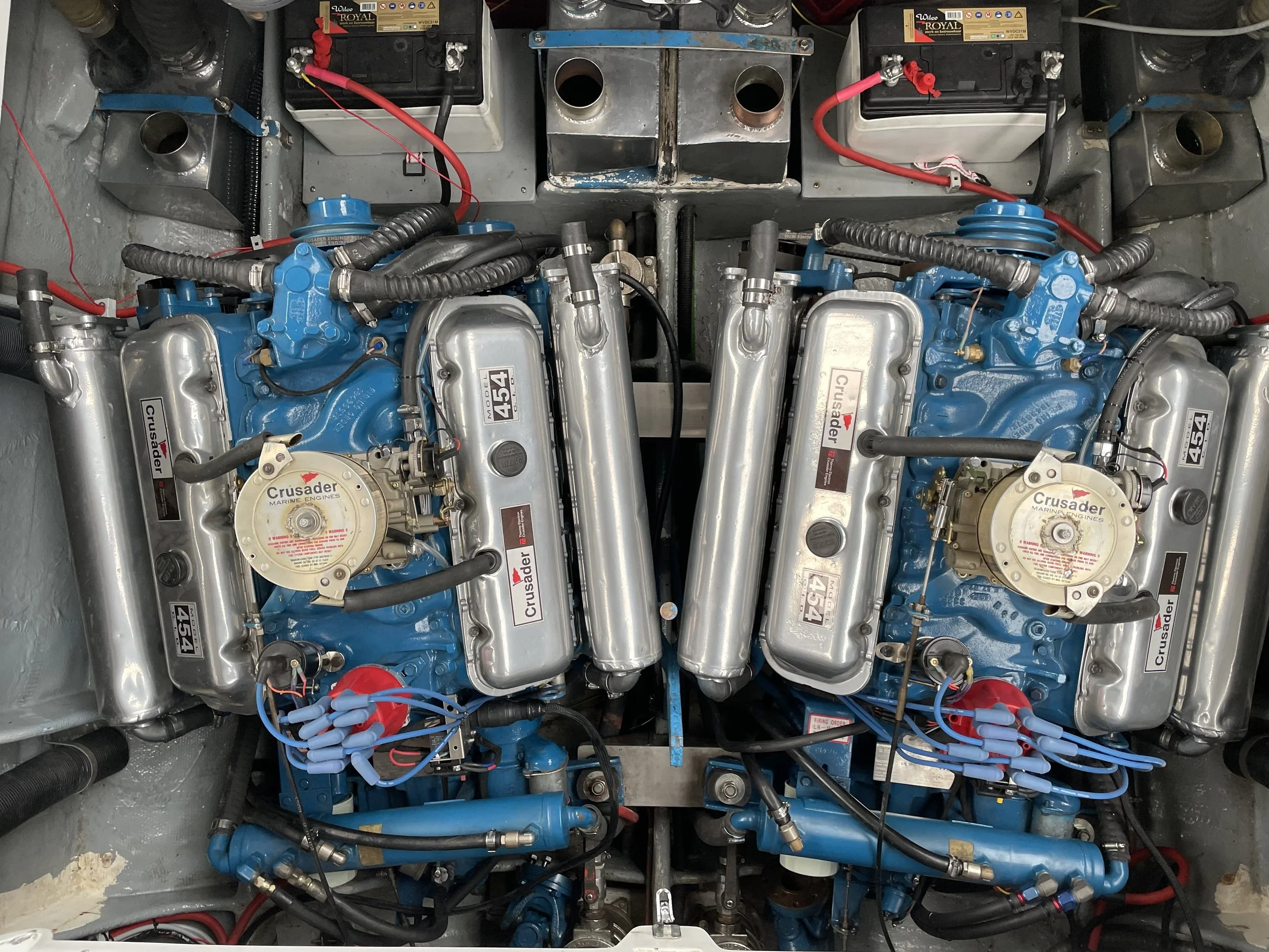 Two blue marine engines with silver components, labeled Crusader, installed in a boat with wiring and hoses around them.