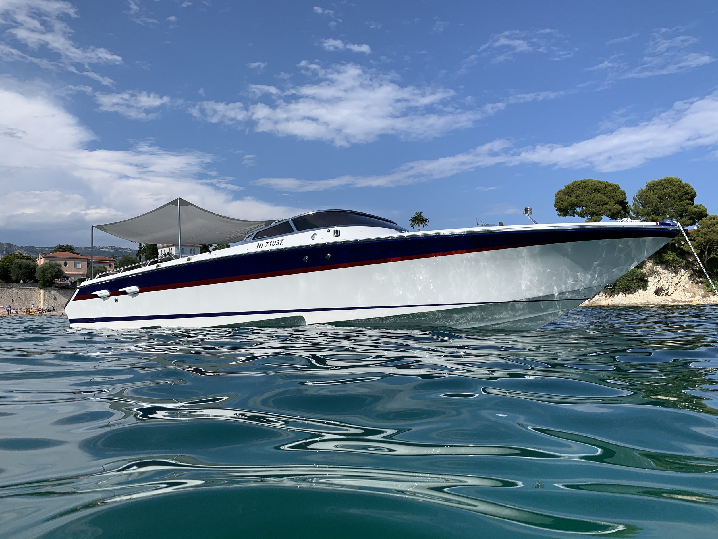 A white yacht with a black and red stripe along the side, floating on calm blue water under a partly cloudy sky, with trees and houses on the shoreline in the background.