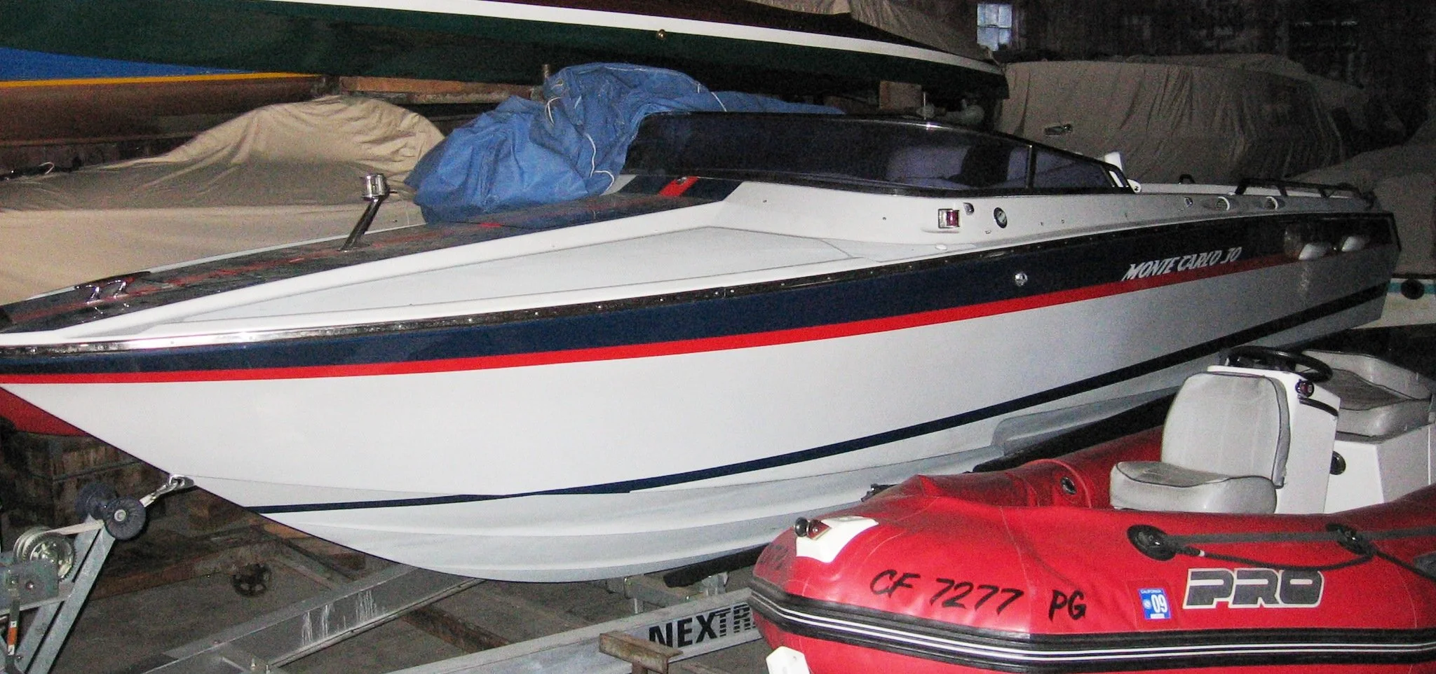 A white racing boat with black and red stripe details, stored on a boat trailer indoors with other boats covered with protective covers around it.