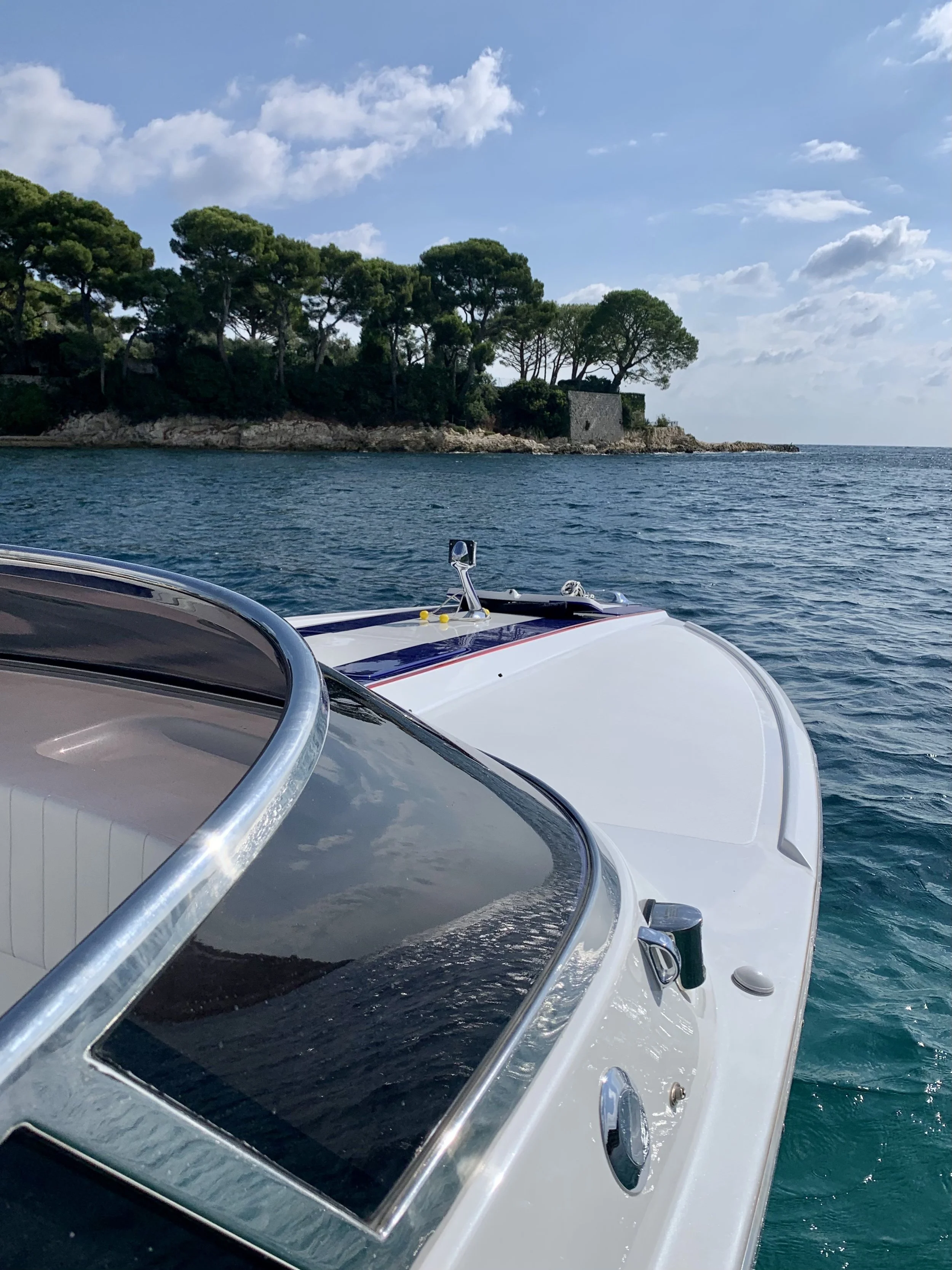 View from a yacht showing the ocean, rocky shoreline with trees, and partially cloudy sky.