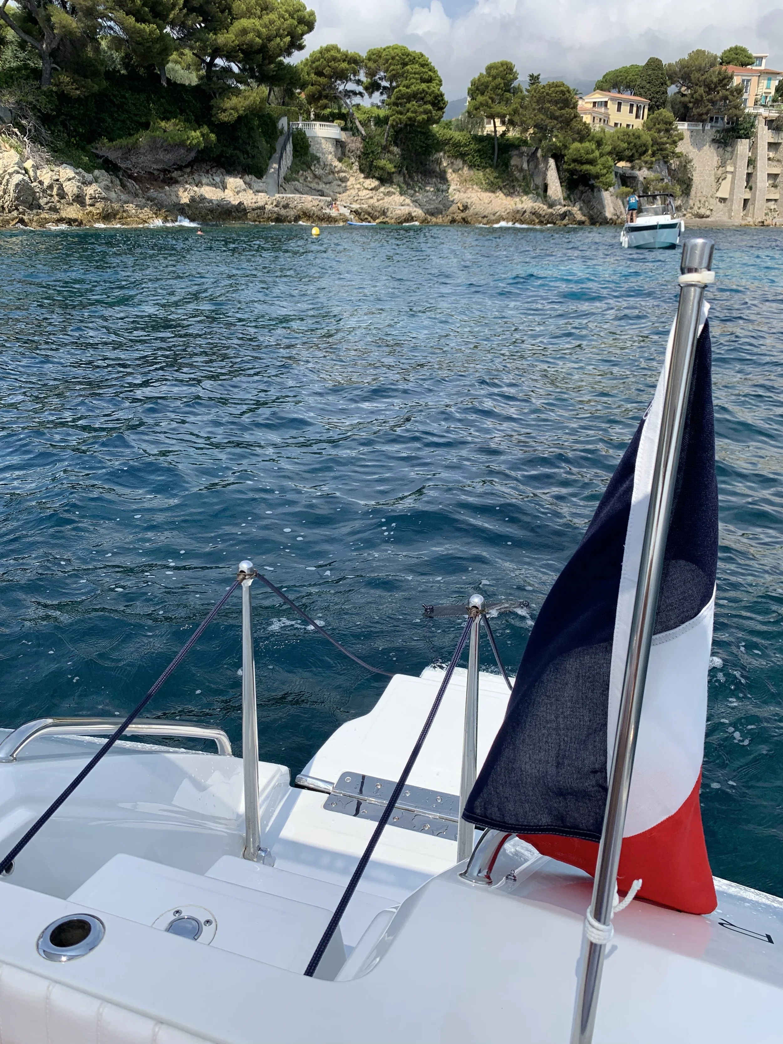 View from the back of a boat on the water with a French flag on a pole. The scene shows a rocky shoreline with trees and some houses in the background, and another boat in the distance.