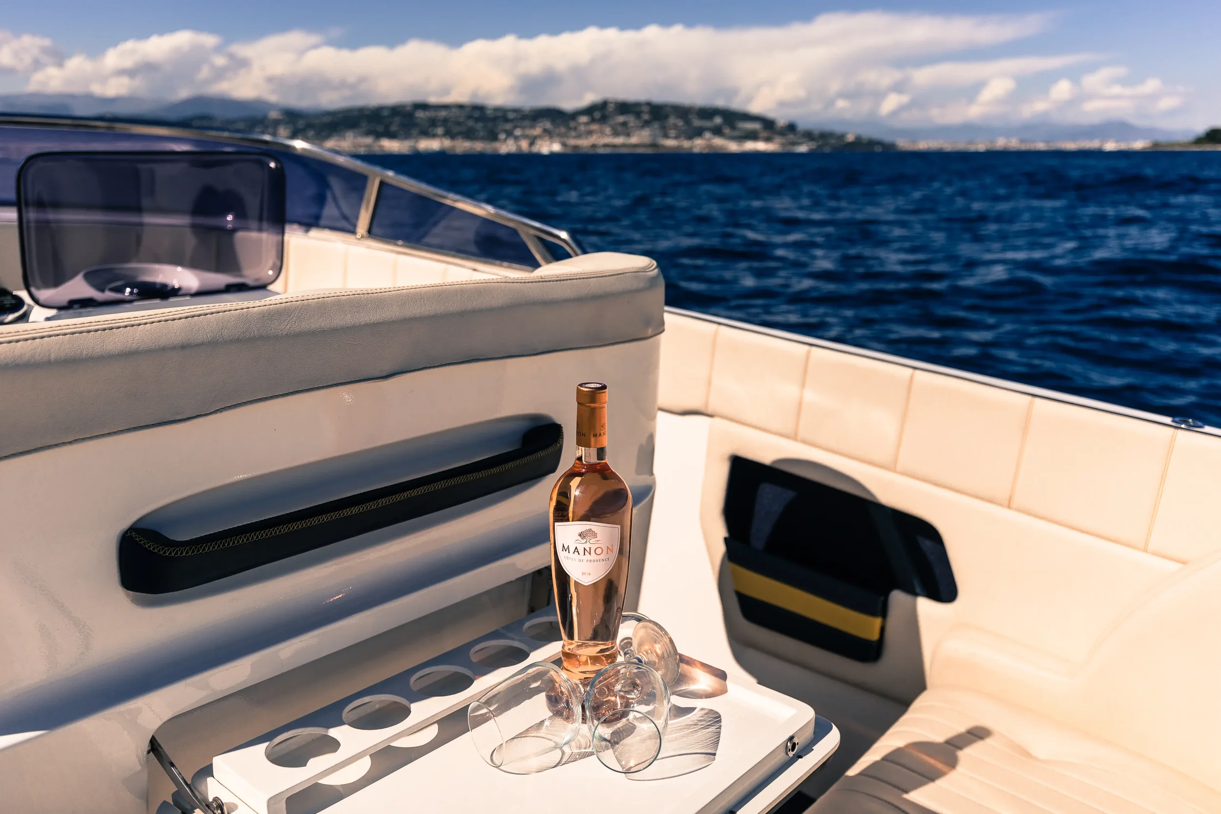 Bottle of Manon rosé wine, three empty wine glasses, on the deck of a yacht with a view of the ocean and distant land and mountains under partly cloudy skies.