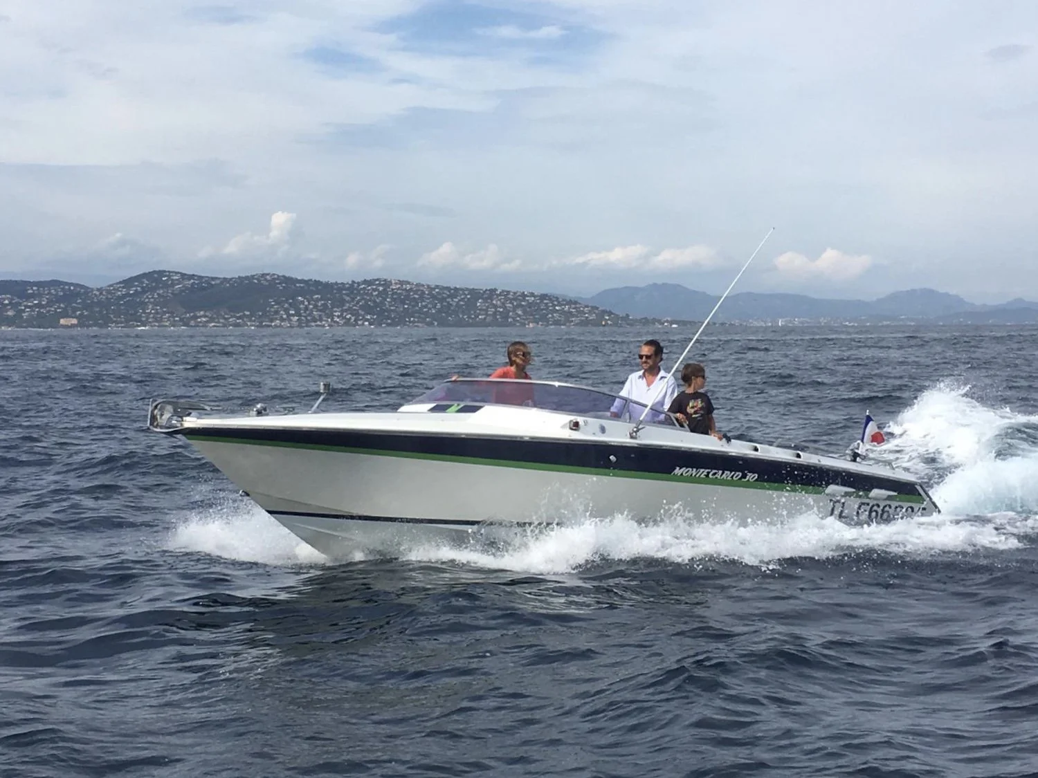 A speedboat with three people on a lake or sea, with a hilly landscape and cloudy sky in the background.