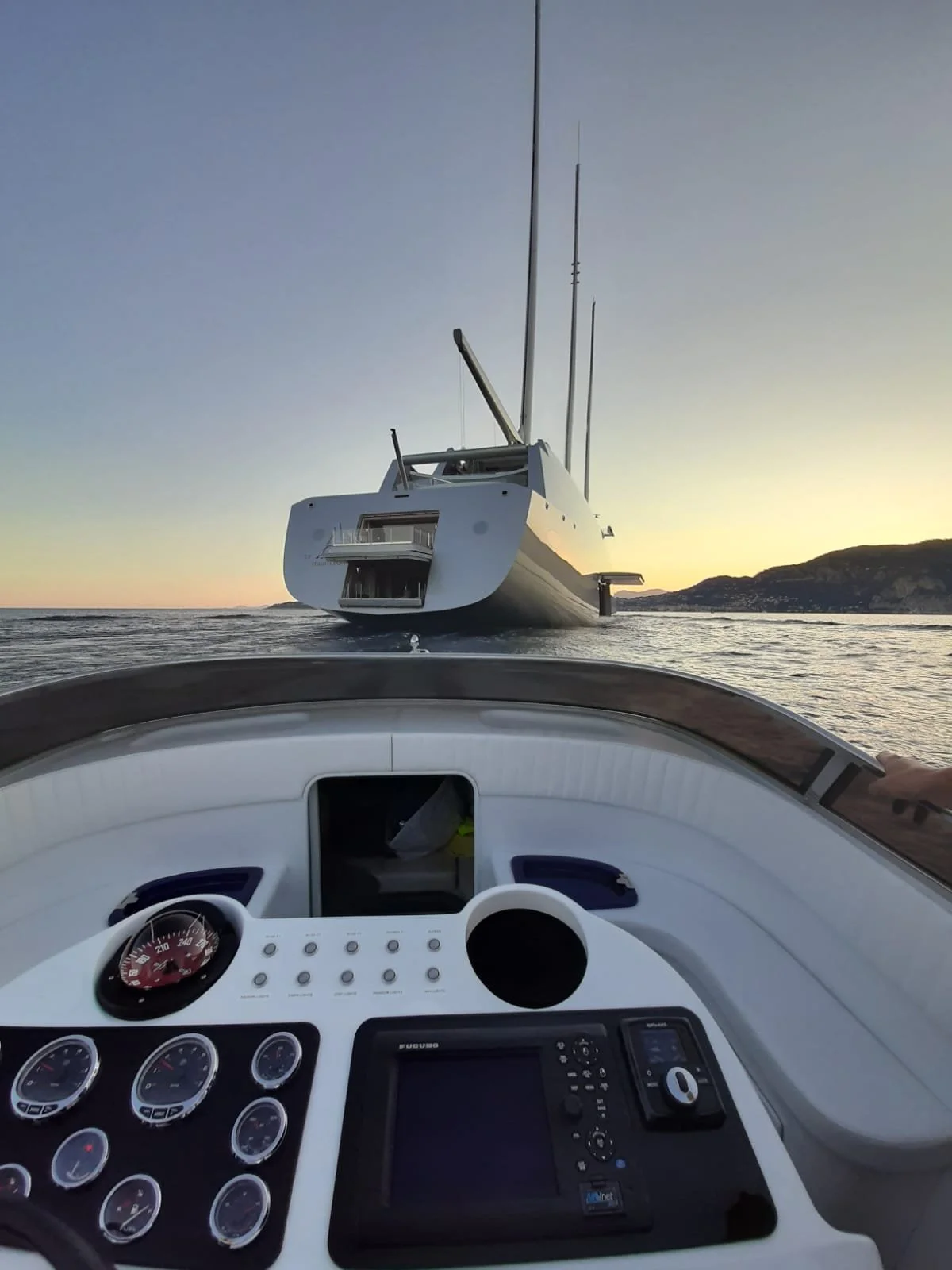 View from the helm of a yacht with a navigation display and gauges, overlooking another yacht on calm water at sunset with a mountain shoreline in the background.