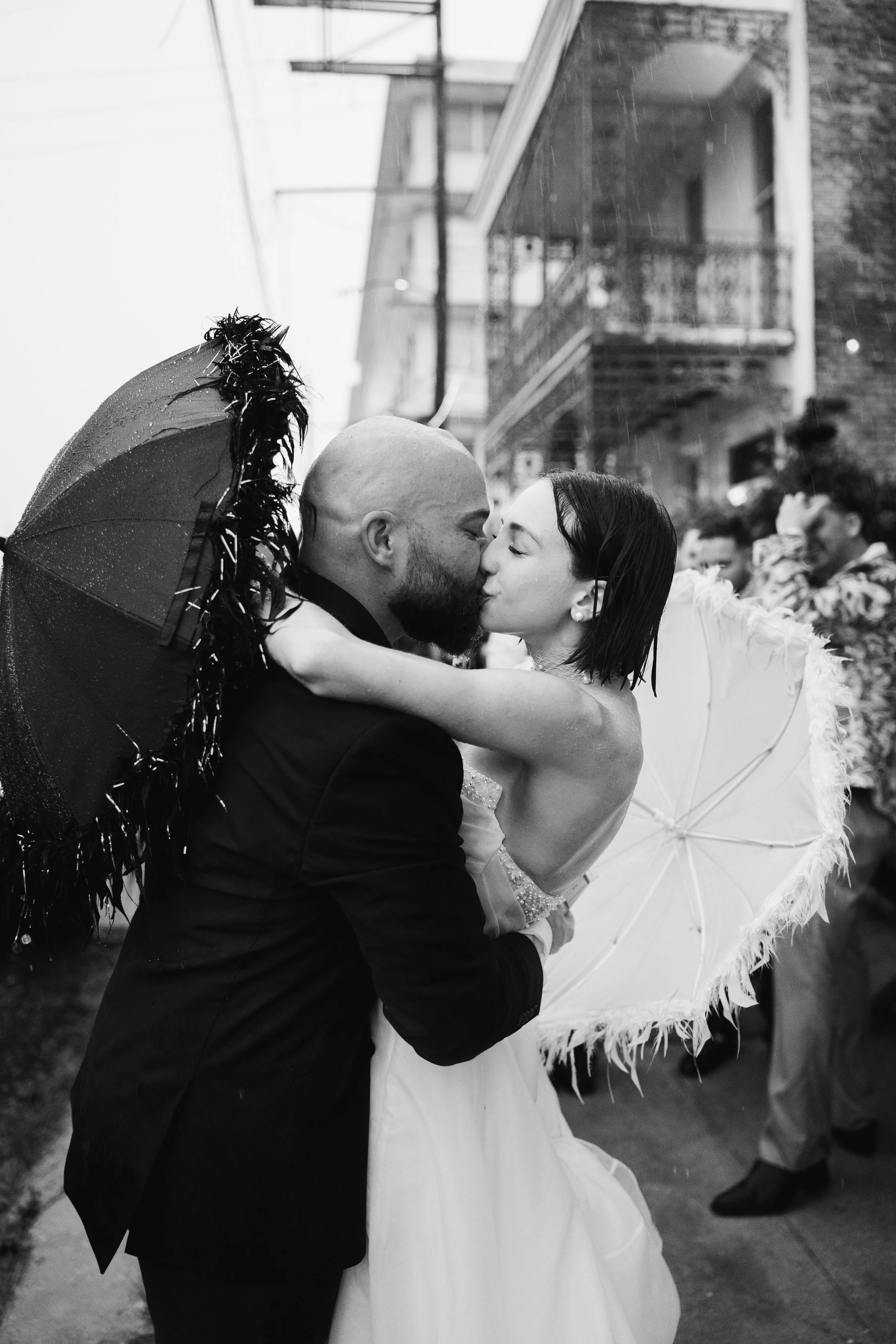 A black and white photograph of a couple sharing a kiss during a rainy outdoor wedding, with the bride holding a white umbrella and the groom holding a decorated umbrella, in a neighborhood setting.