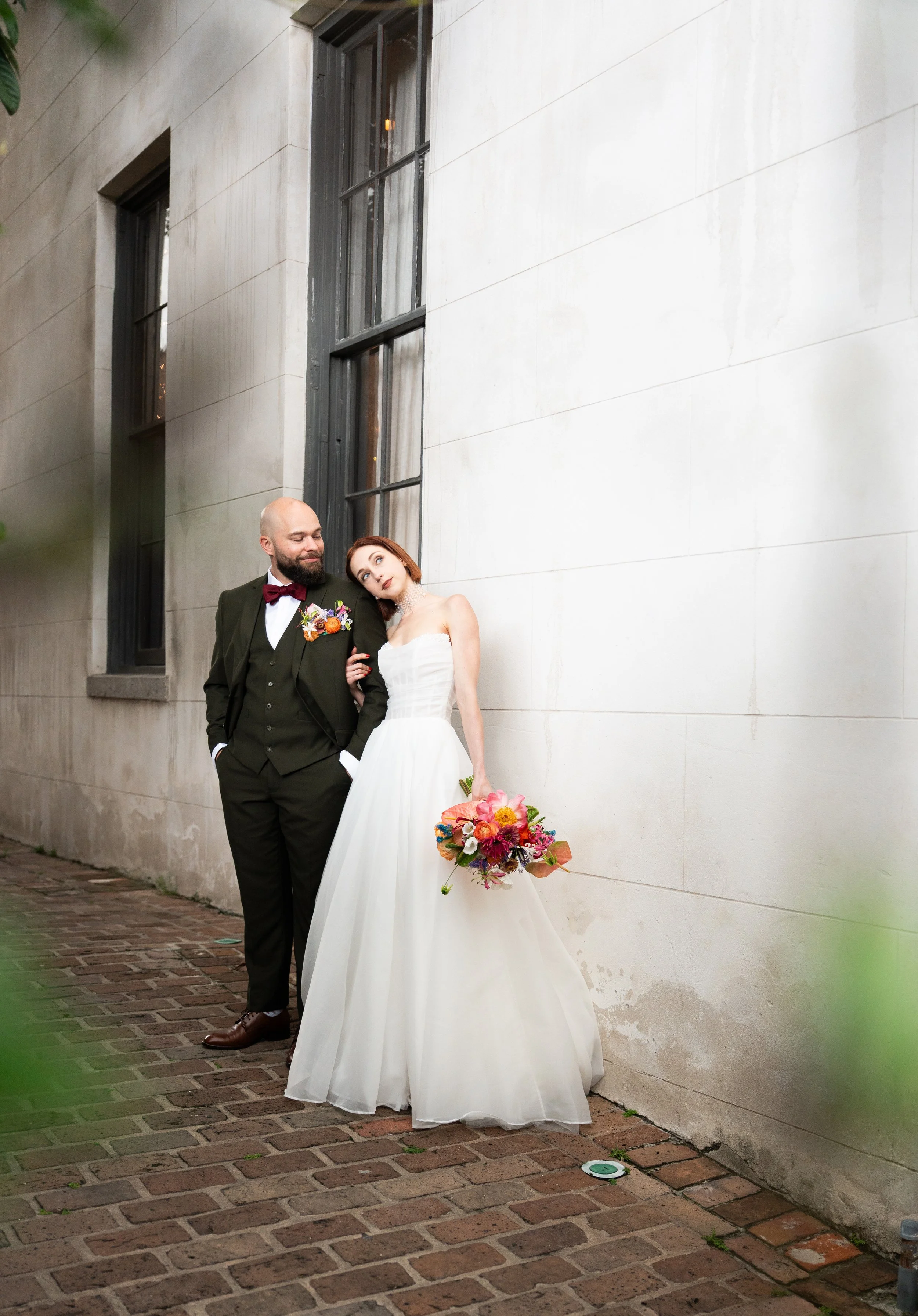 A bride and groom standing together outside near a wall, with the bride holding a colorful bouquet and leaning her head on the groom's shoulder, both dressed in wedding attire.