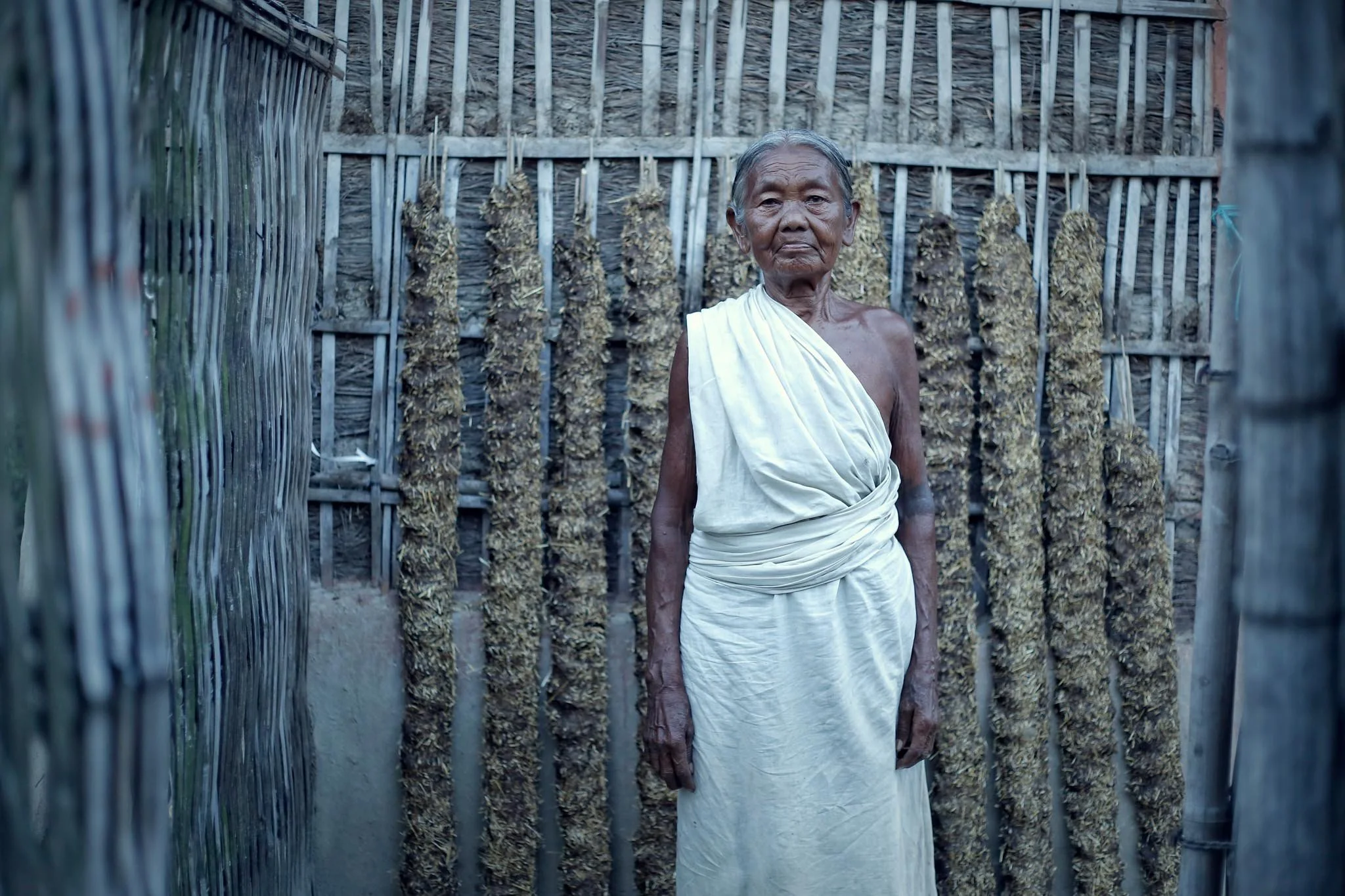 An elderly man in traditional white clothing standing inside a bamboo and grass hut with hanging plants. Sustainable living of the Tharu community in Nepal. A project for Doner Agencies by photographer Gautam Dhimal