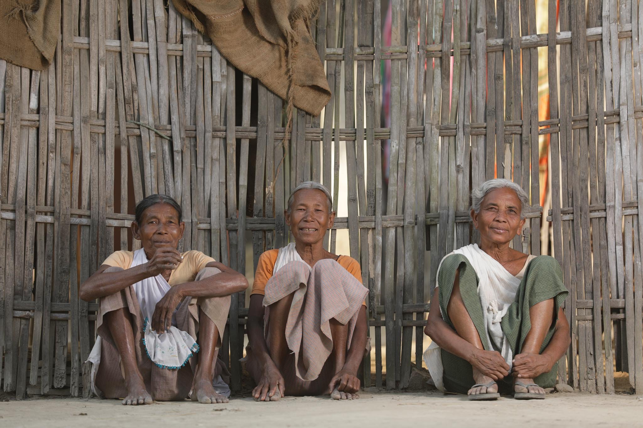 Three elderly Tharu women sitting cross-legged in front of a woven bamboo wall, with relaxed expressions. Photography by Gautam Dhimal in Sunsari, Nepal