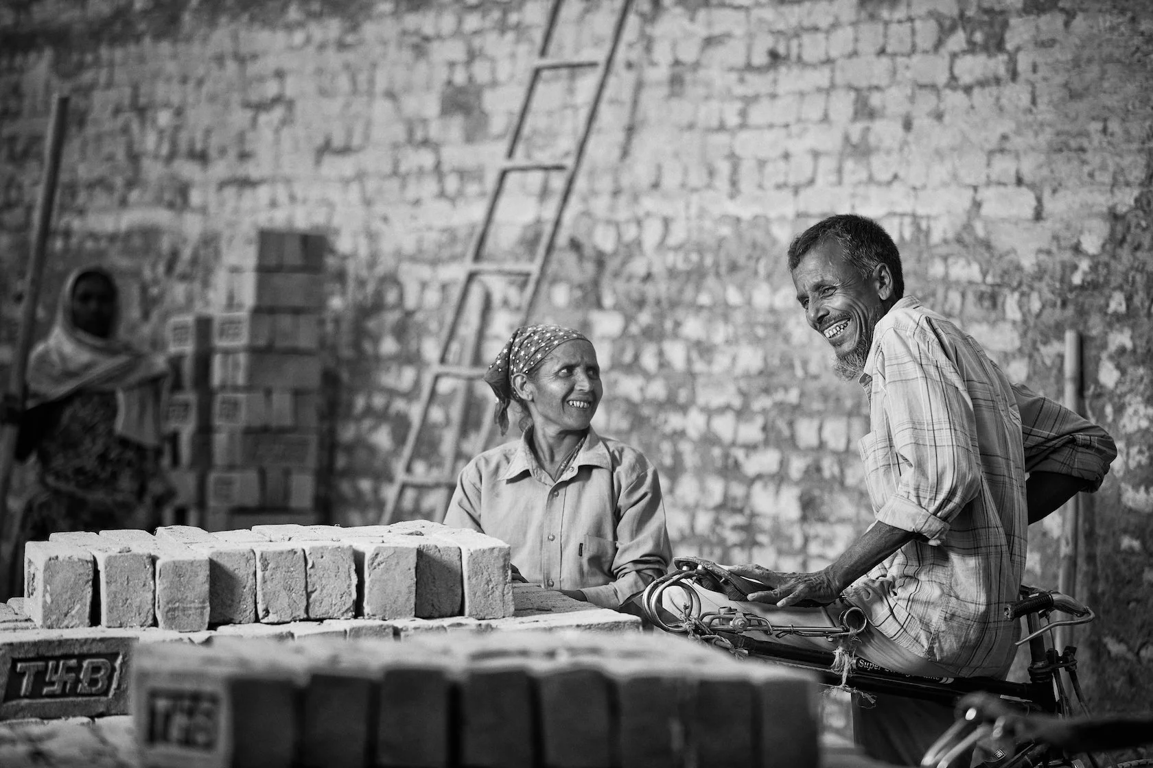 Two elderly people, a man and a woman, smiling and talking while sitting on a bicycle, surrounded by bricks and construction supplies in front of a brick wall.