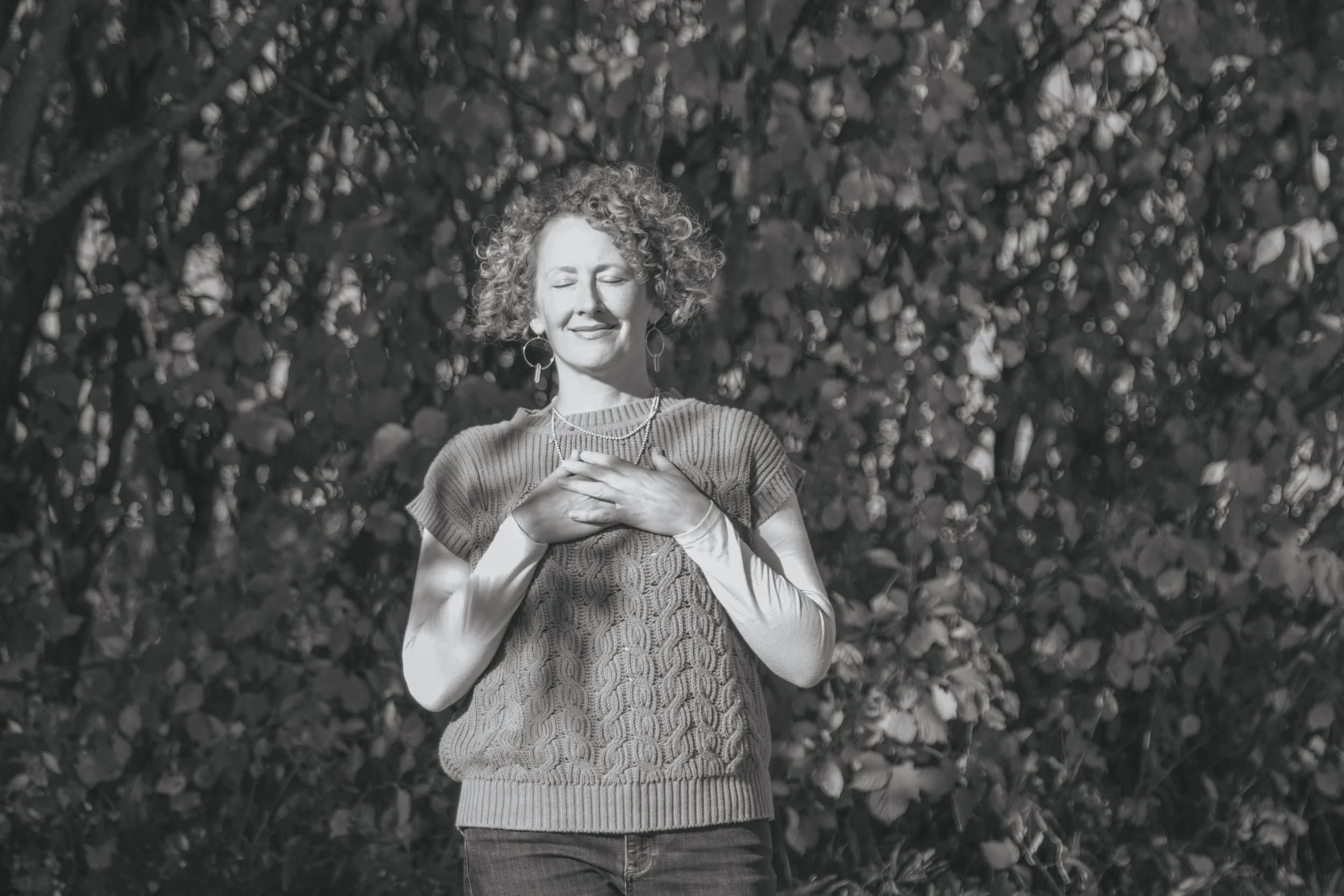 a black and white photo of a curly haired woman wearing a long sleeved shirt under a sleeveless sweater with her hands over her heart