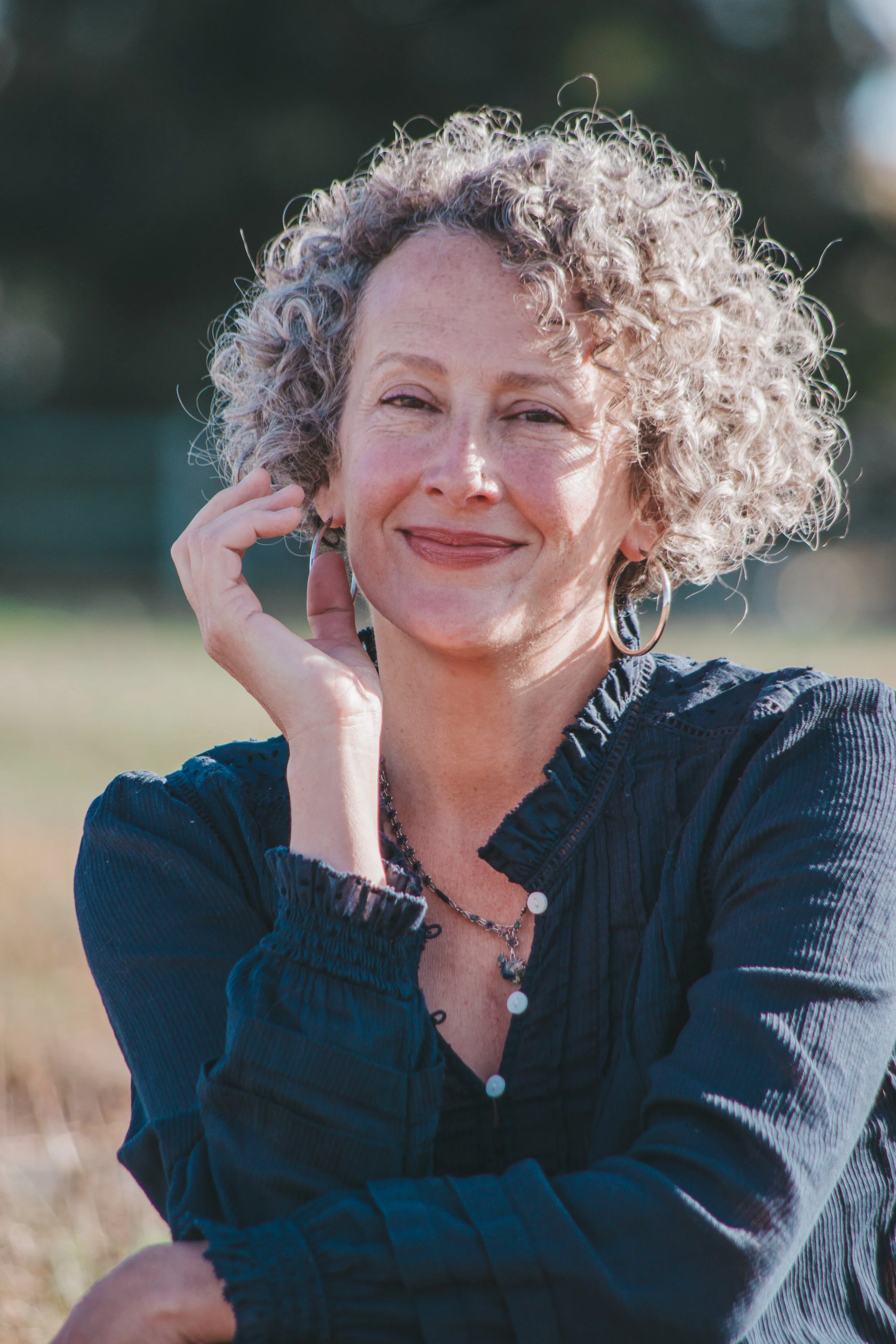 a fair skinned woman with a curly grey lob wearing a navy blouse gesture with her hand near her face sitting in a field