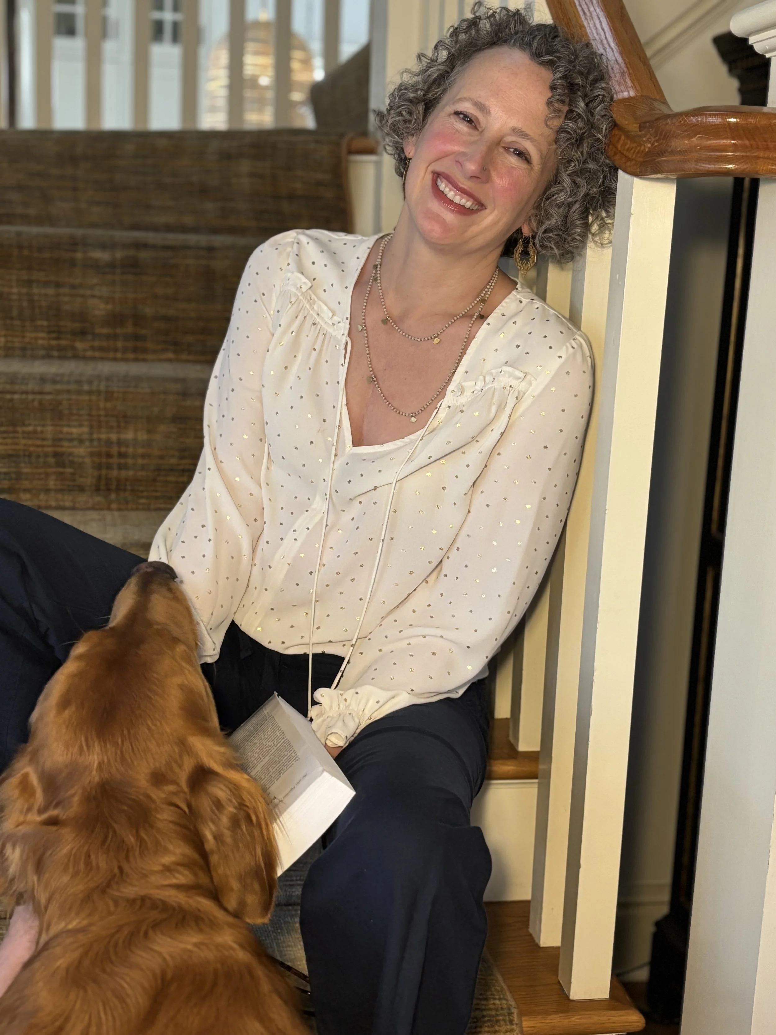 a grey & curly haired woman wearing a cream colored v-neck blouse leans against a banister with a book in one hand and a golden retriever in the foreground