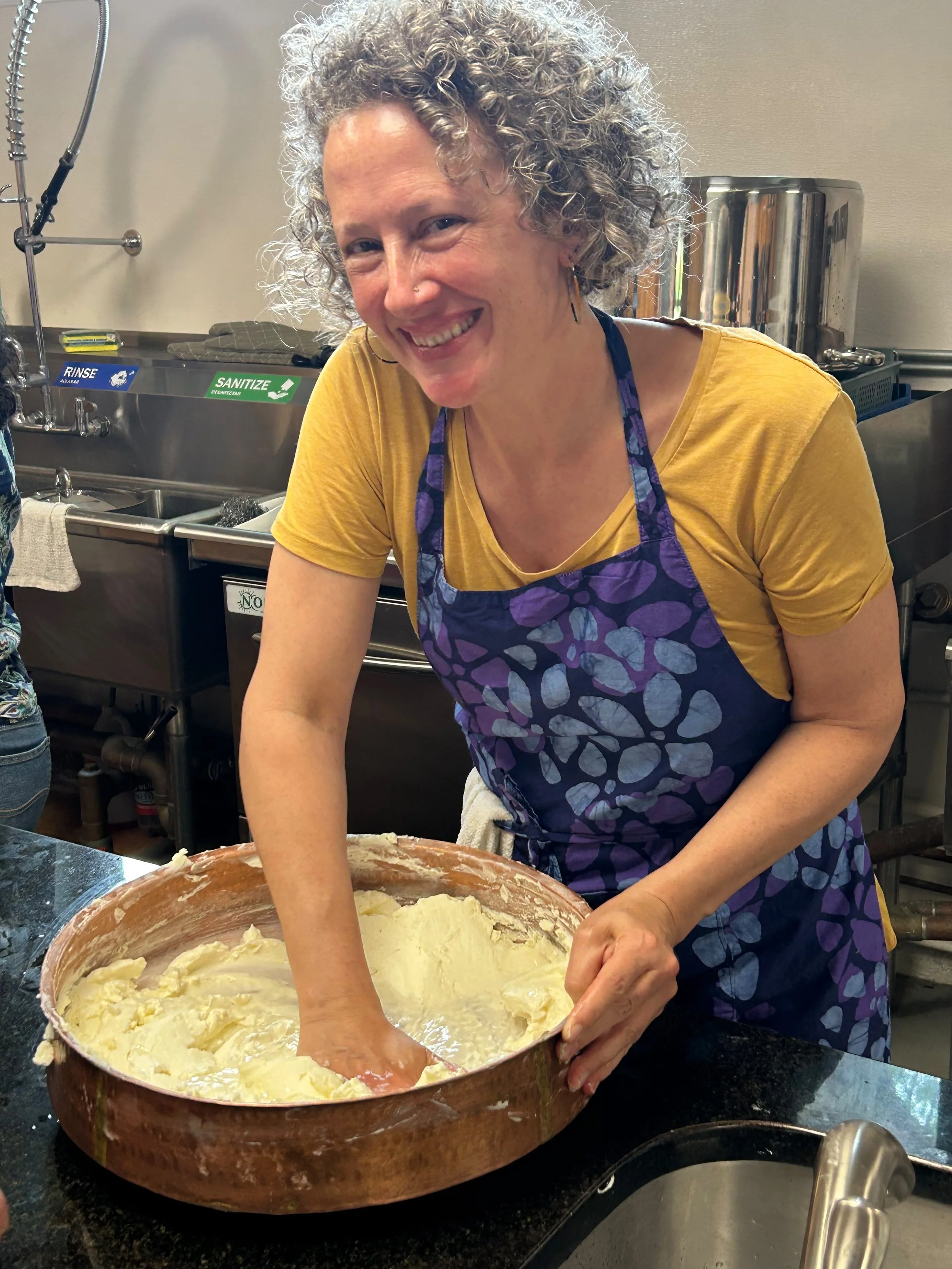 wearing a yellow t shirt and blue patterned apron, standing in a commercial kitchen, Kimberly hand-stirs ghee in a large copper bowl.