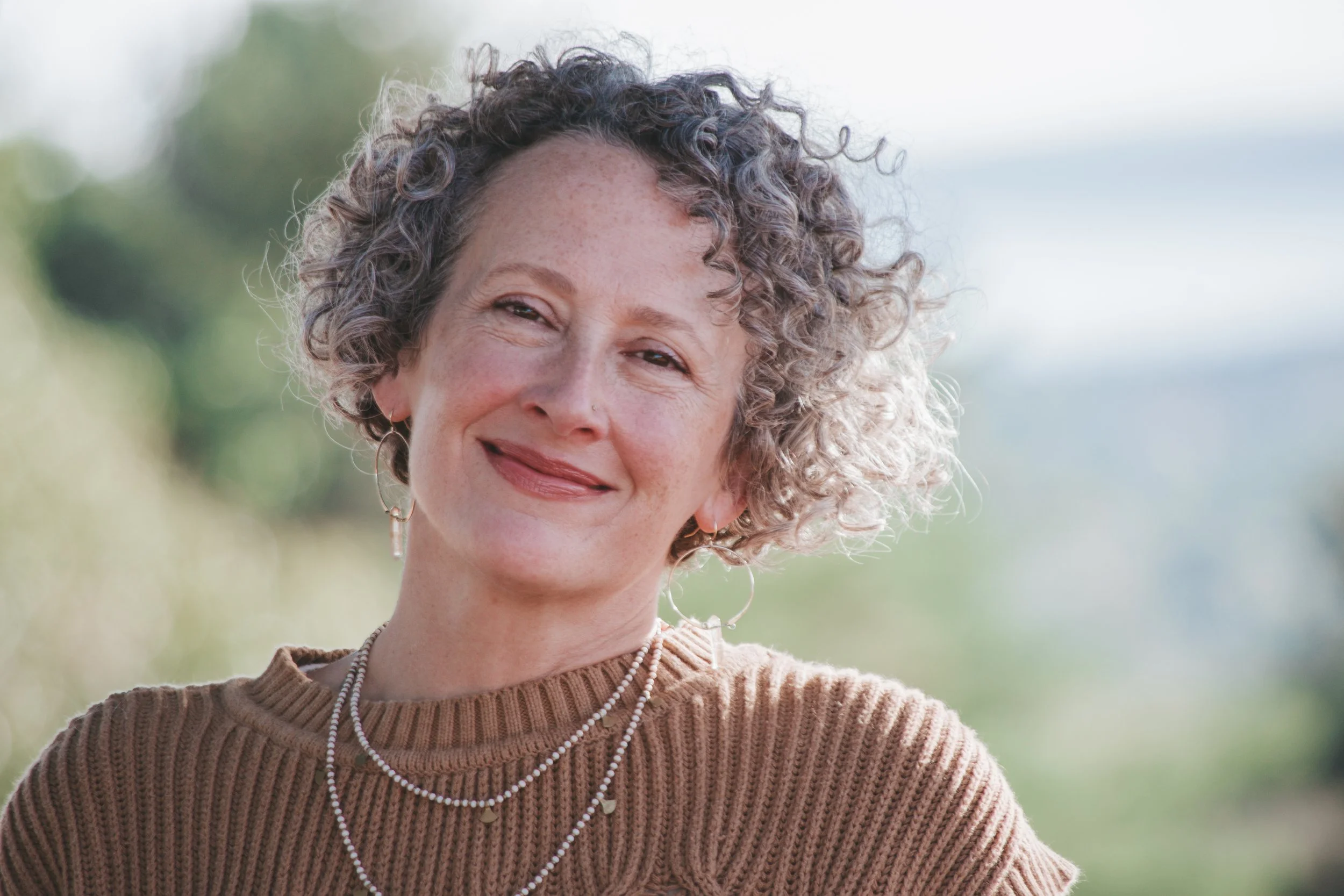 Kimberly wearing a dark amber sweater, a double strand of white beads and gold hoops with citrine crystals, offers a gentle smile to the viewer. The background is blurry but bright outdoor environment.