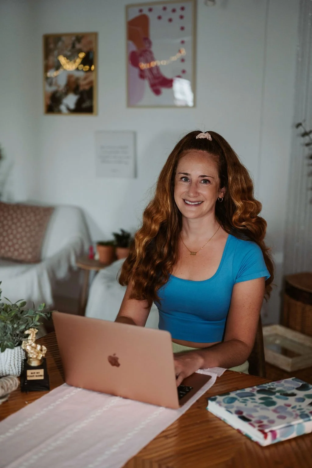 A woman with long, curly red hair smiling while sitting at a wooden table with a pink laptop in front of her, in a cozy, well-decorated room.