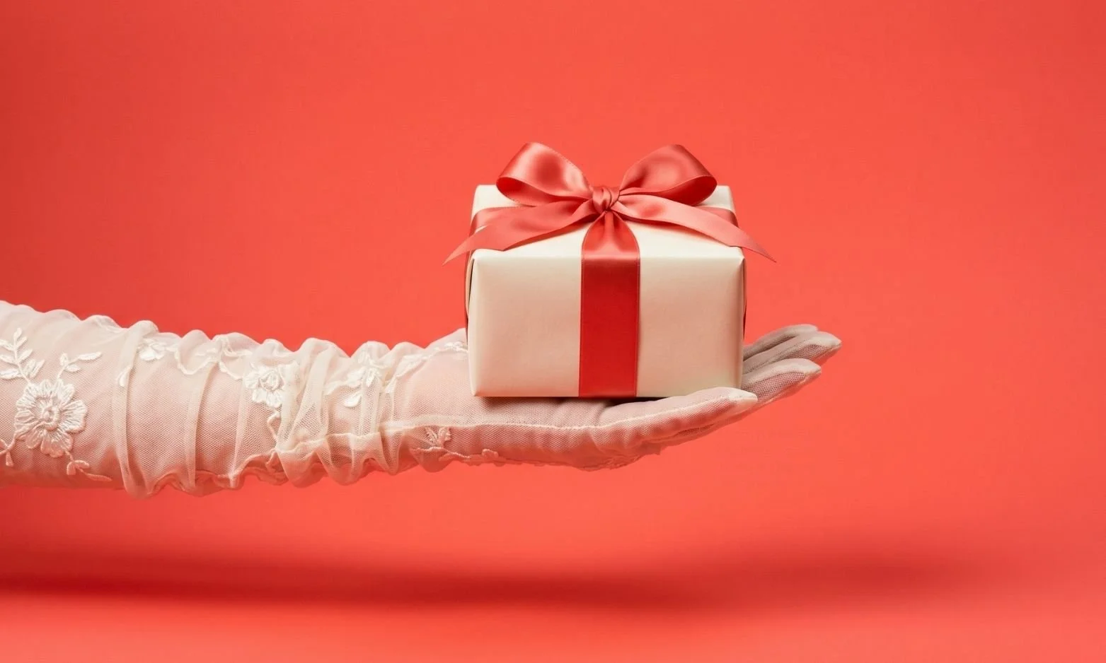 Ivory tulle-gloved hand offering a small gift-wrapped package on a coral-red infinity cove, representing the zero price effect and how the concept of "free" triggers distinct psychological responses compared to low prices