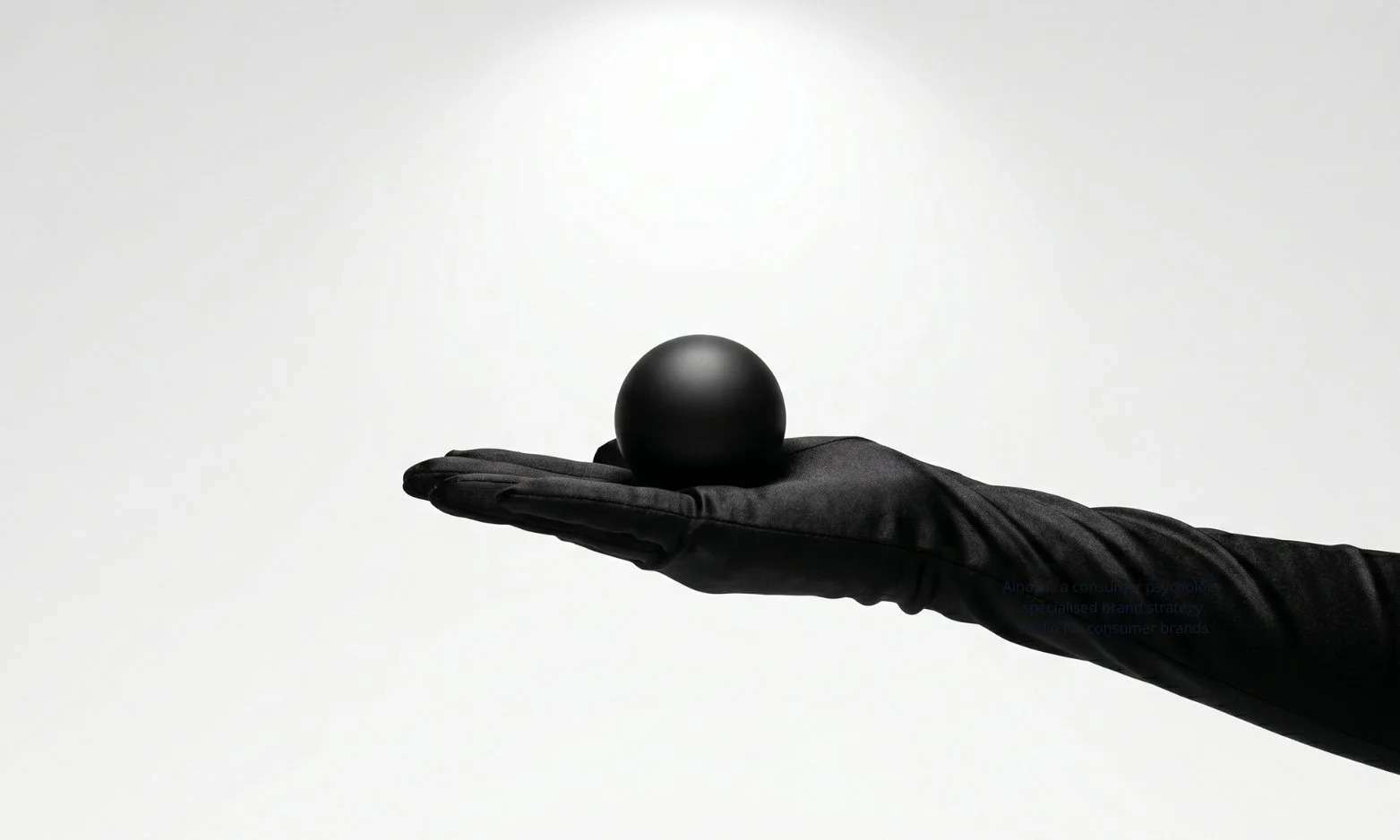 Black satin-gloved hand presenting a matte black sphere on a white infinity cove — representing prestige pricing and the psychological link between high price, restraint, and perceived quality.