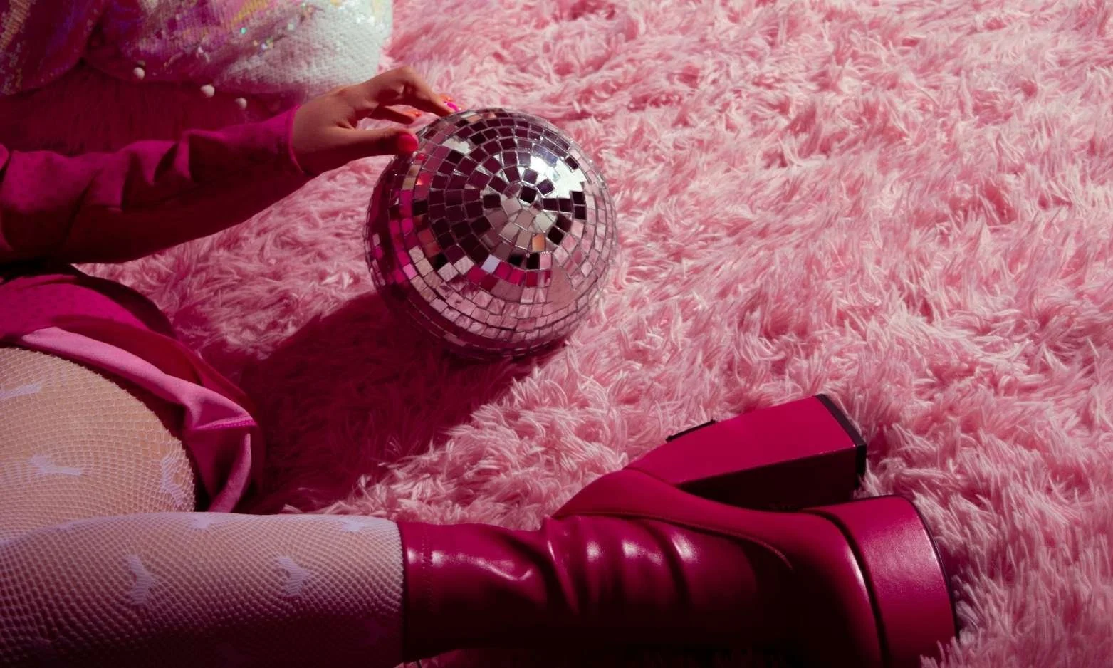 Bratz aesthetic: woman in pink leopard coloured dress, pink heart fish net stockings, thick pink leather ankle boots with thick heel, holding a disco ball lightly with pink nails while laying on pinky fluffy rug.