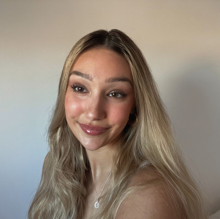 Close-up of a young woman with long blonde hair smiling at the camera against a plain wall.