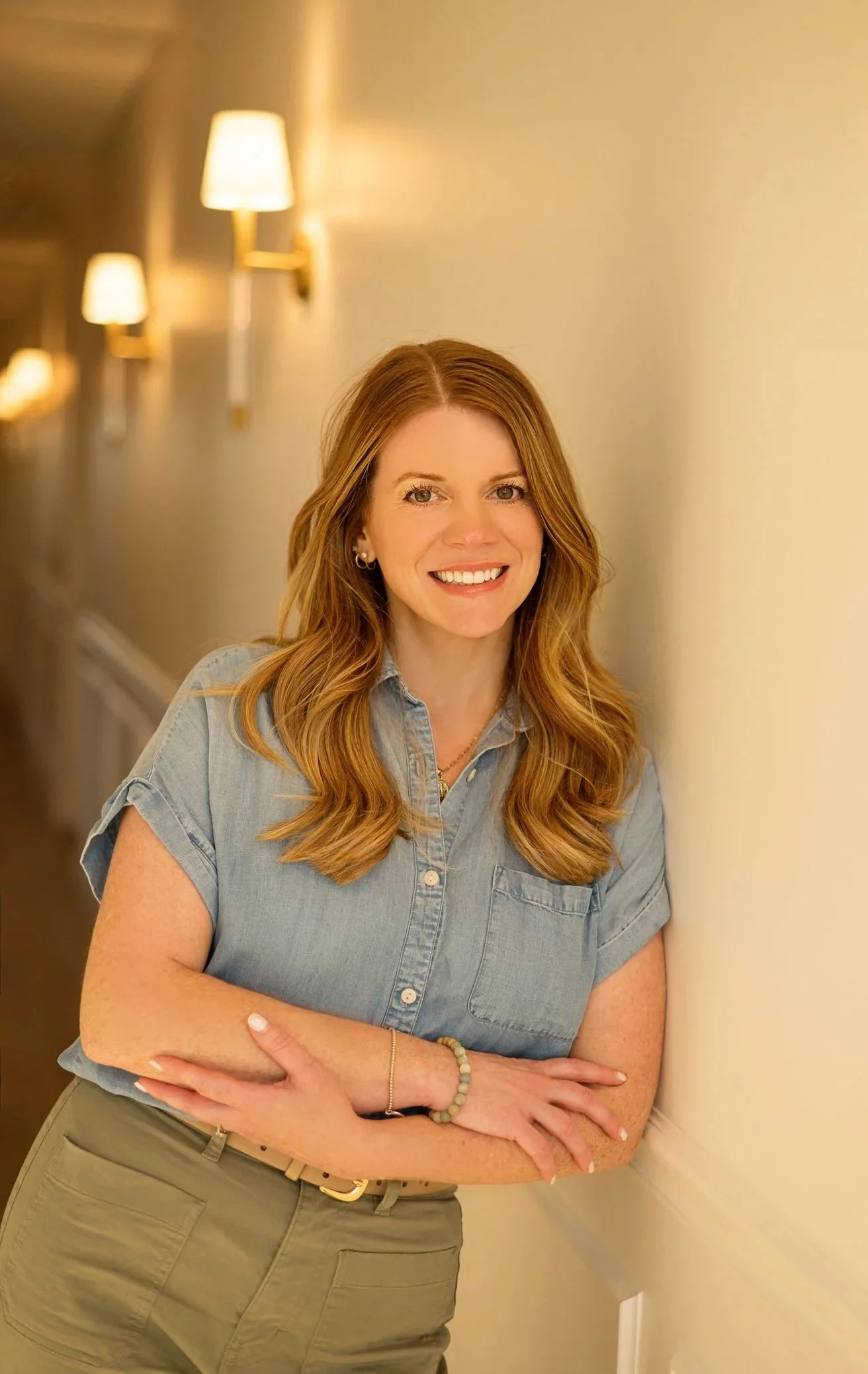A smiling woman with long red hair standing against a beige wall with lights along a hallway.
