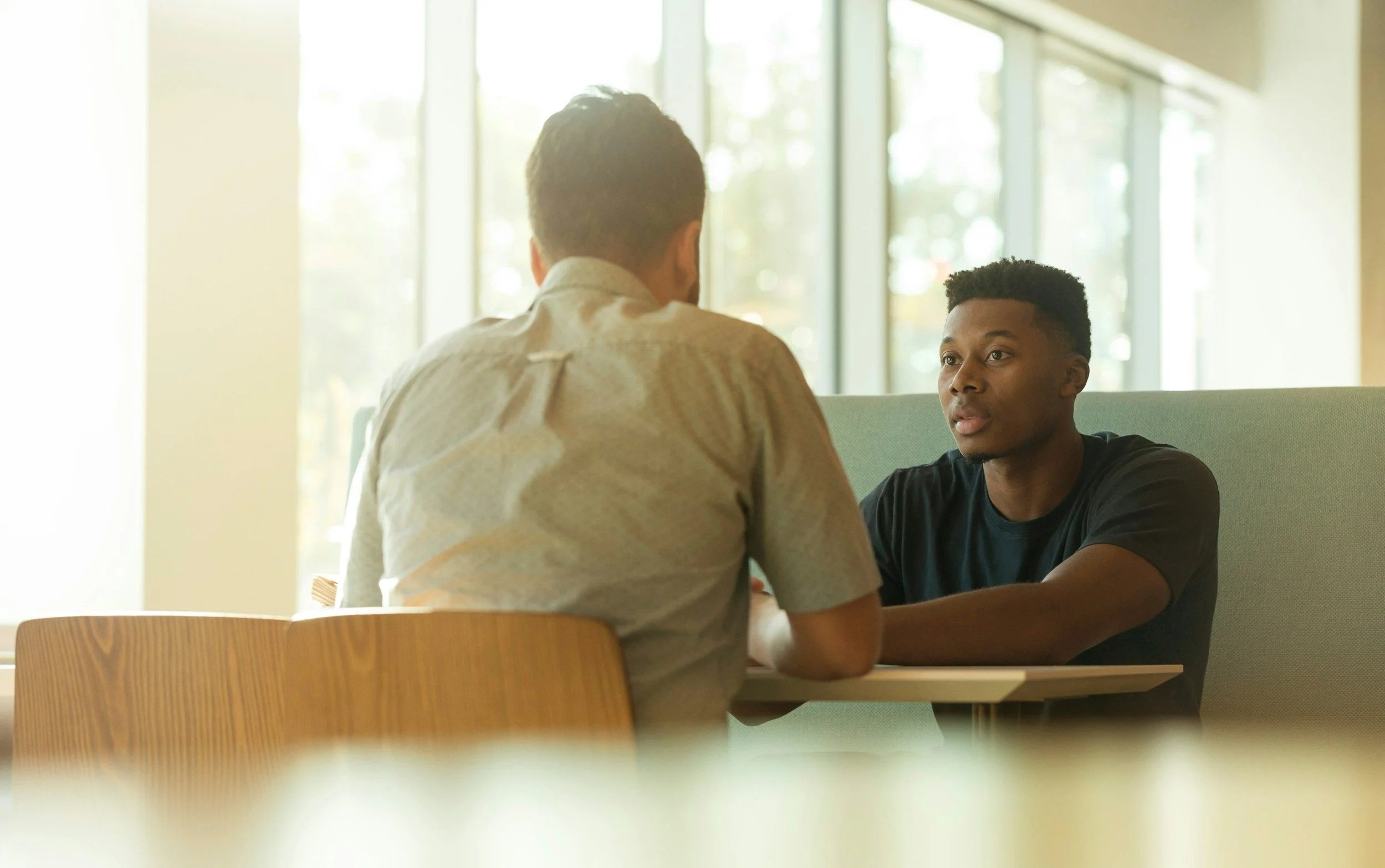 Two men sitting at a table having a conversation in a well-lit room with large windows.