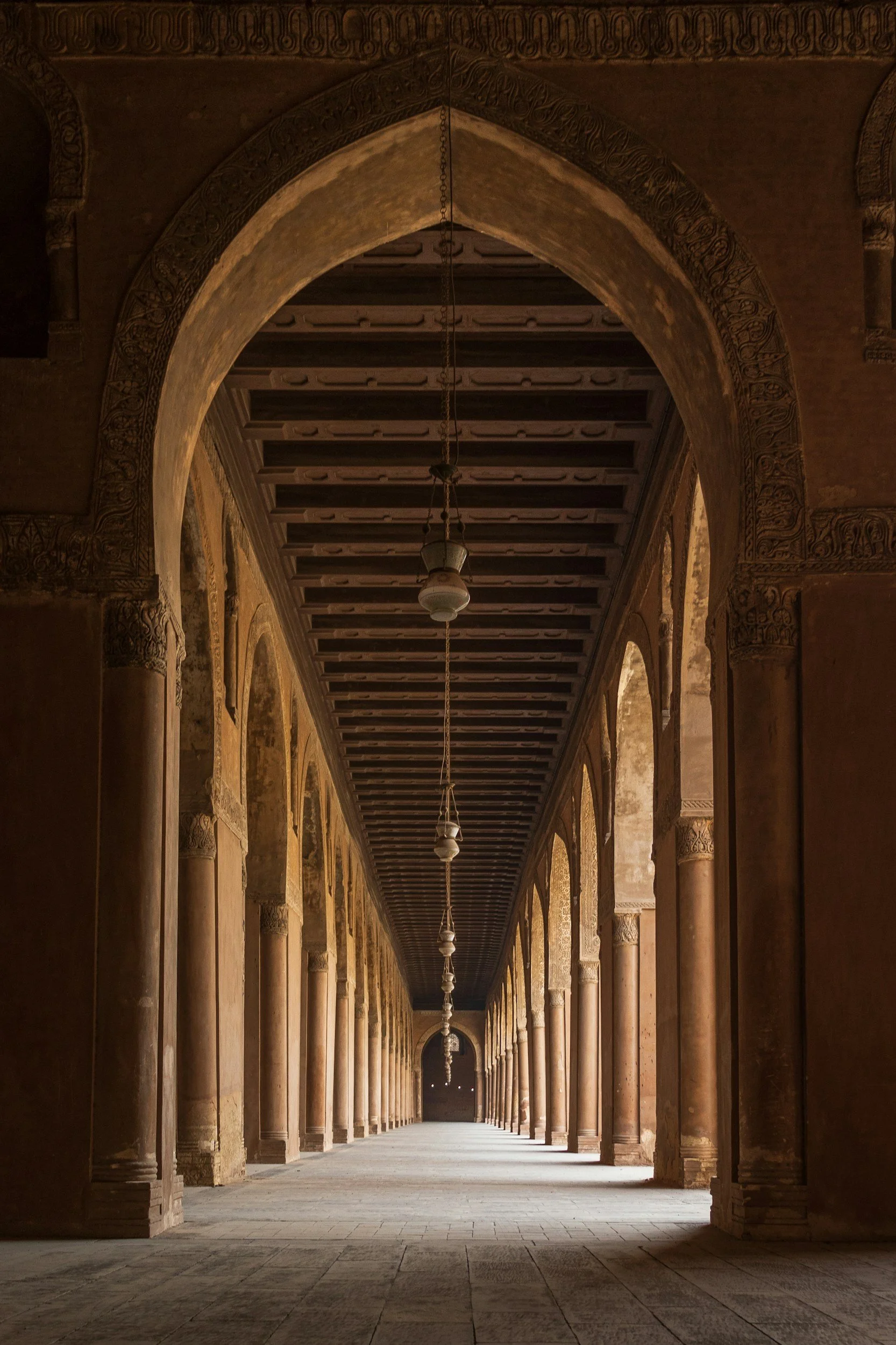 A long corridor with high arches and columns, ornate detailing on the arches, and hanging light fixtures, inside an ancient historic building.