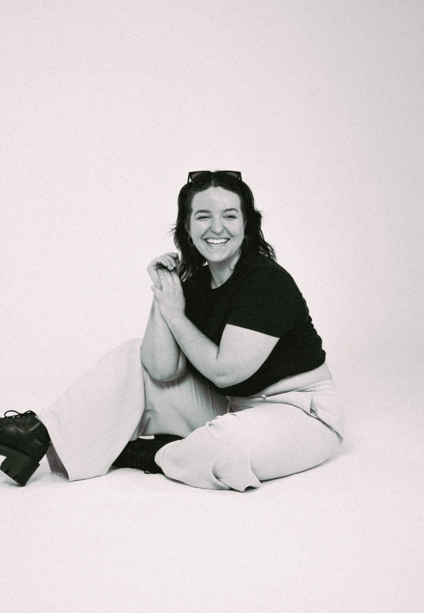 A young woman with dark hair, sunglasses on her head, wearing a black t-shirt and light-colored pants, sitting on the floor against a plain light background, smiling.