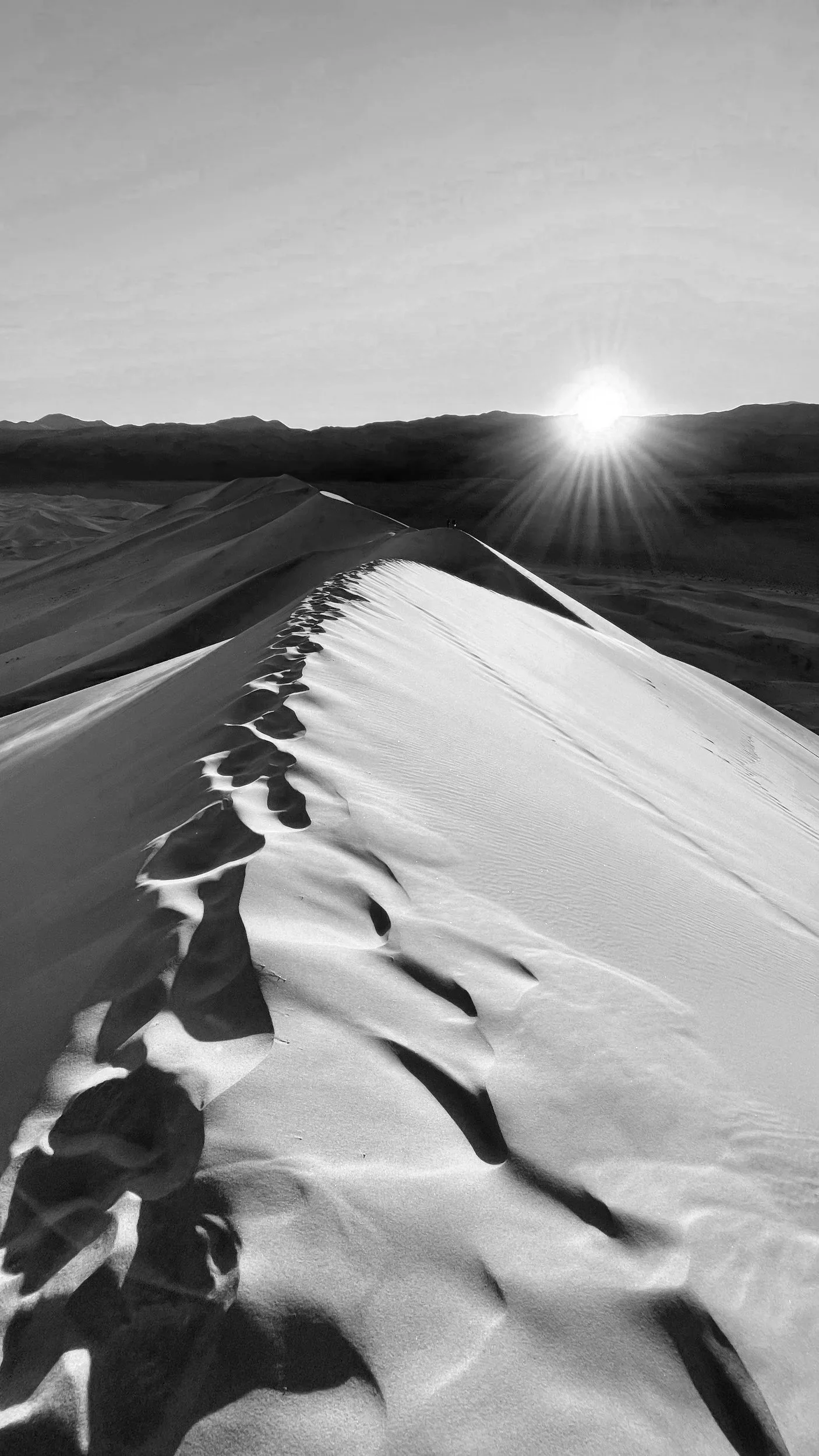 Black and white photo of a sandy desert dune with footprints leading up the slope, and the sun setting behind distant mountains.