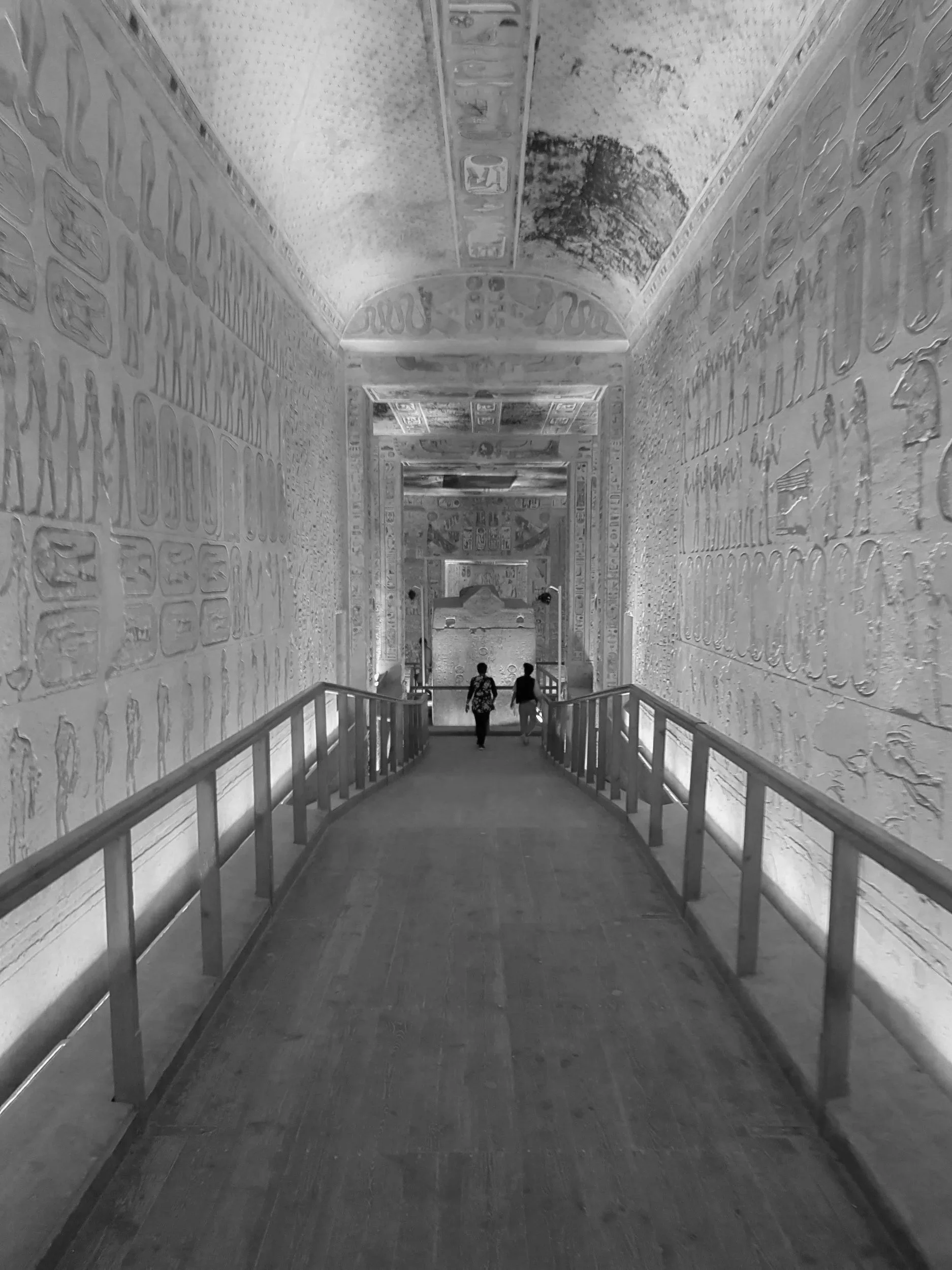 Two people walk down an ancient passageway with hieroglyphics and artwork on the walls and ceiling, in a historical site, in black and white.