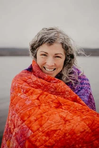 Smiling woman with curly gray hair wrapped in a colorful quilt outdoors near water.