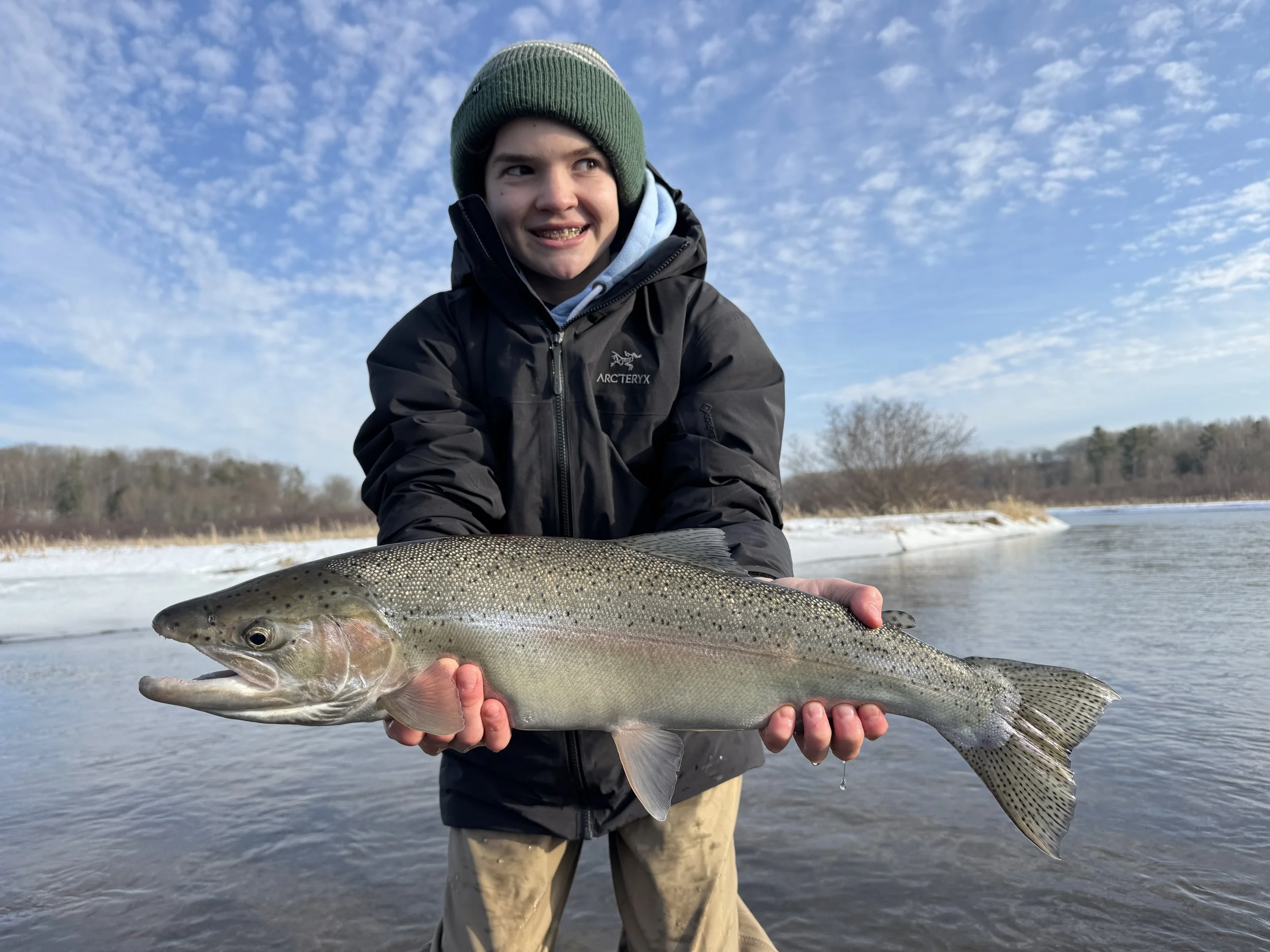 A boy dressed in winter clothing and a green beanie holding a large rainbow trout in front of a river and winter landscape.