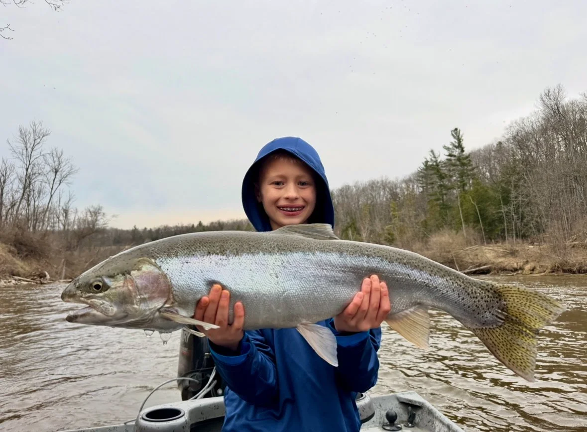 A young boy smiling while holding a large fish he caught in a river, with leafless trees in the background under an overcast sky.