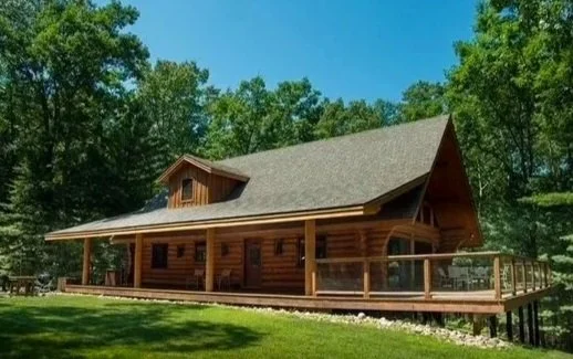 Wooden cabin with a porch and railing, surrounded by trees and grass under a clear blue sky.