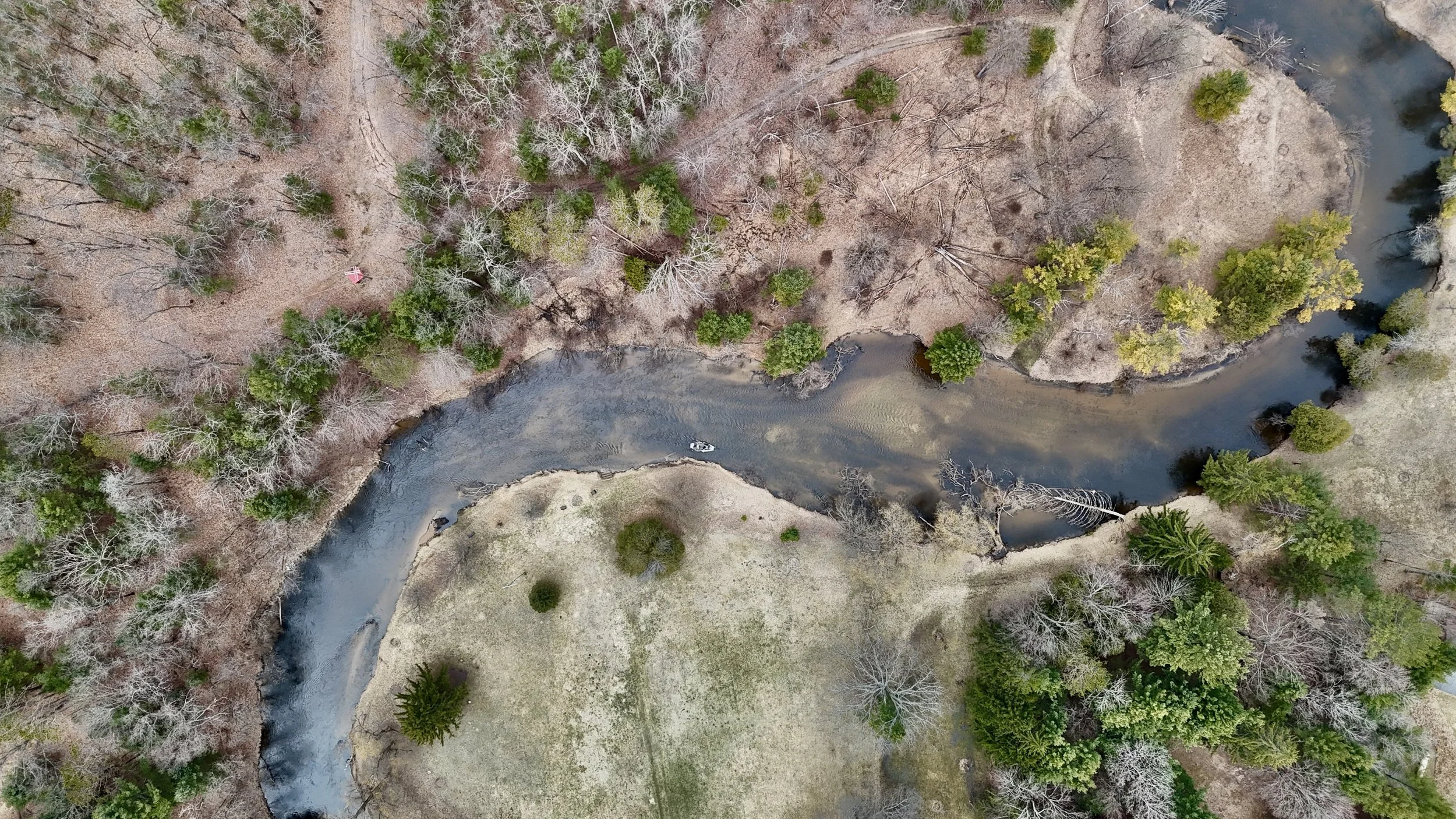 Aerial view of a winding river flowing through a landscape with leafless trees and some evergreen trees, with grassy areas and a boat in the water.