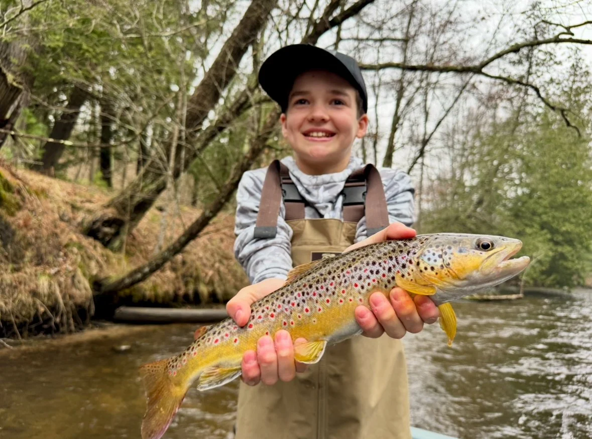 A boy wearing overalls and a cap, standing in a creek holding a large rainbow trout with a smile on his face.