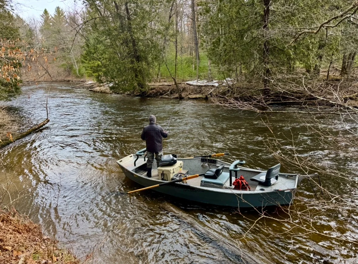 A person standing in a small boat on a narrow river surrounded by trees with some new spring leaves.