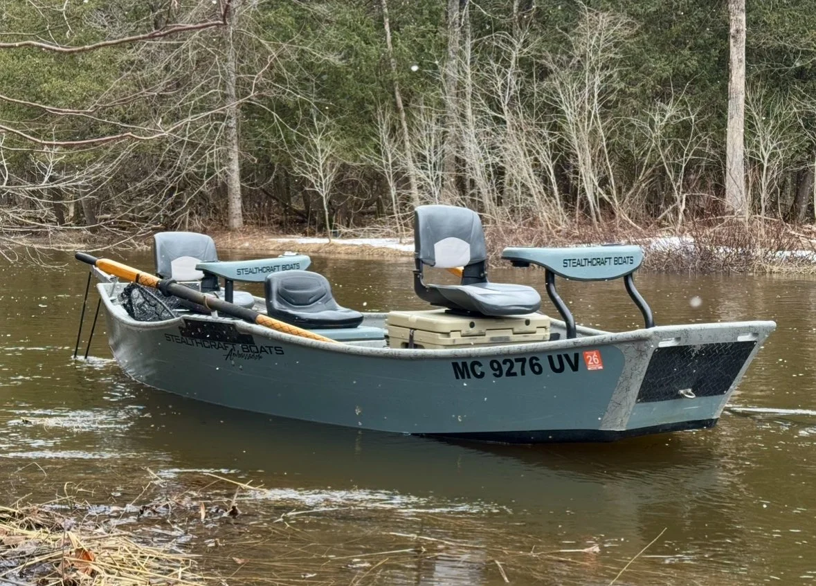 A gray fishing boat on a river with four gray seats, an oar, and trees in the background.