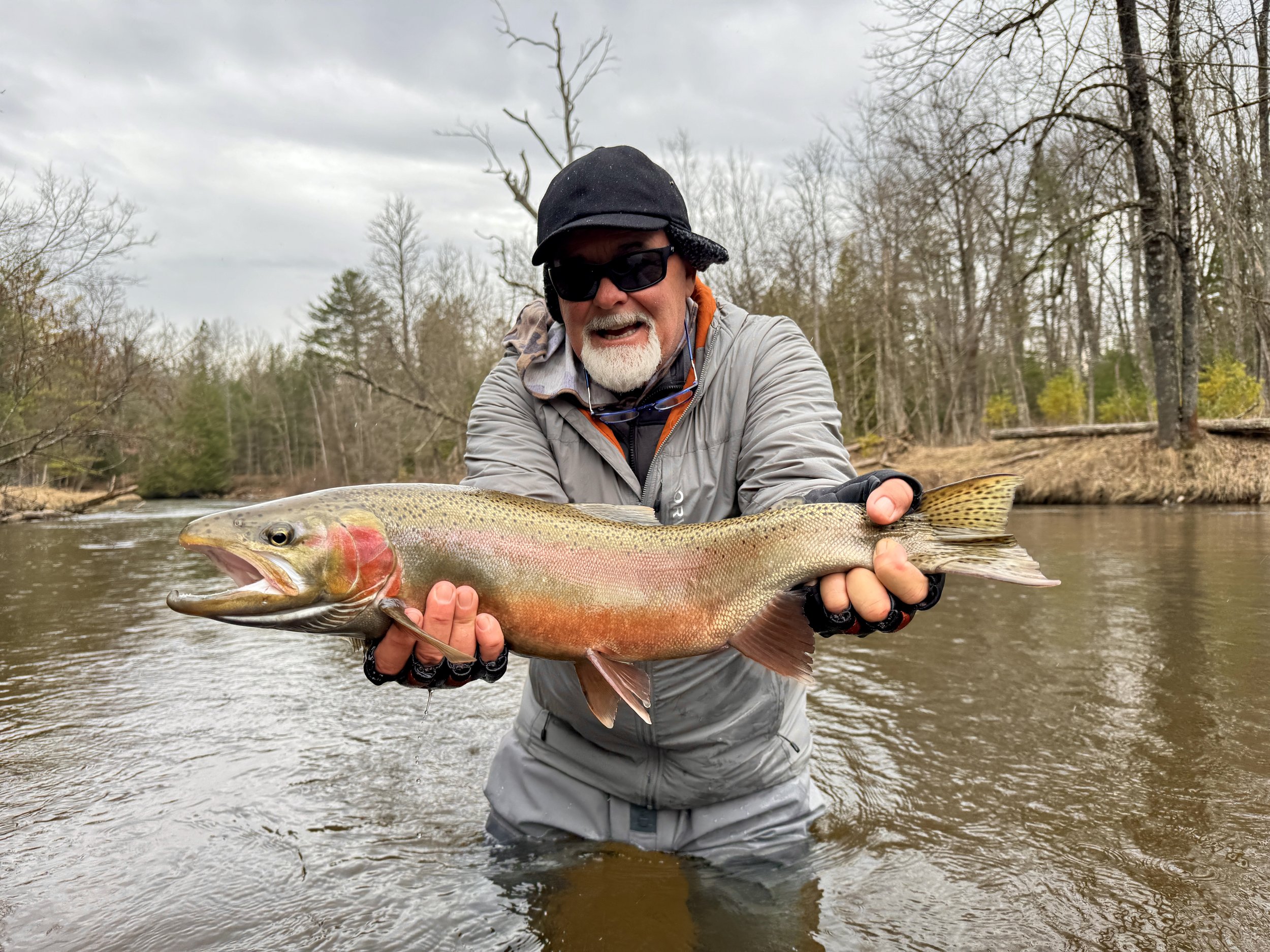 A man wearing a black hat, sunglasses, and a gray jacket standing in a river, holding a large rainbow trout with both hands.