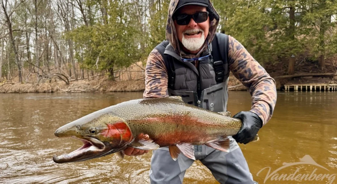 A man wearing sunglasses, a hooded jacket, and gloves is standing in a river, holding a large rainbow trout he caught.